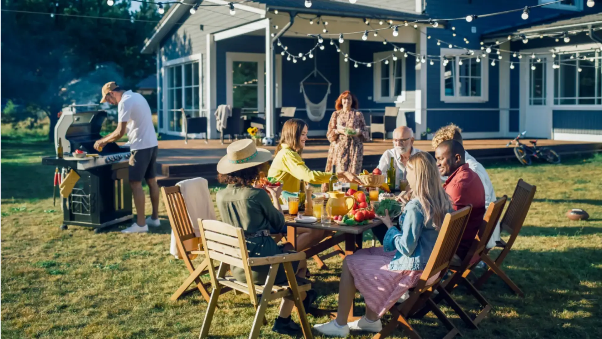 Family enjoying backyard housewarming party with barbecue, outdoor dining, and string lights at cozy home