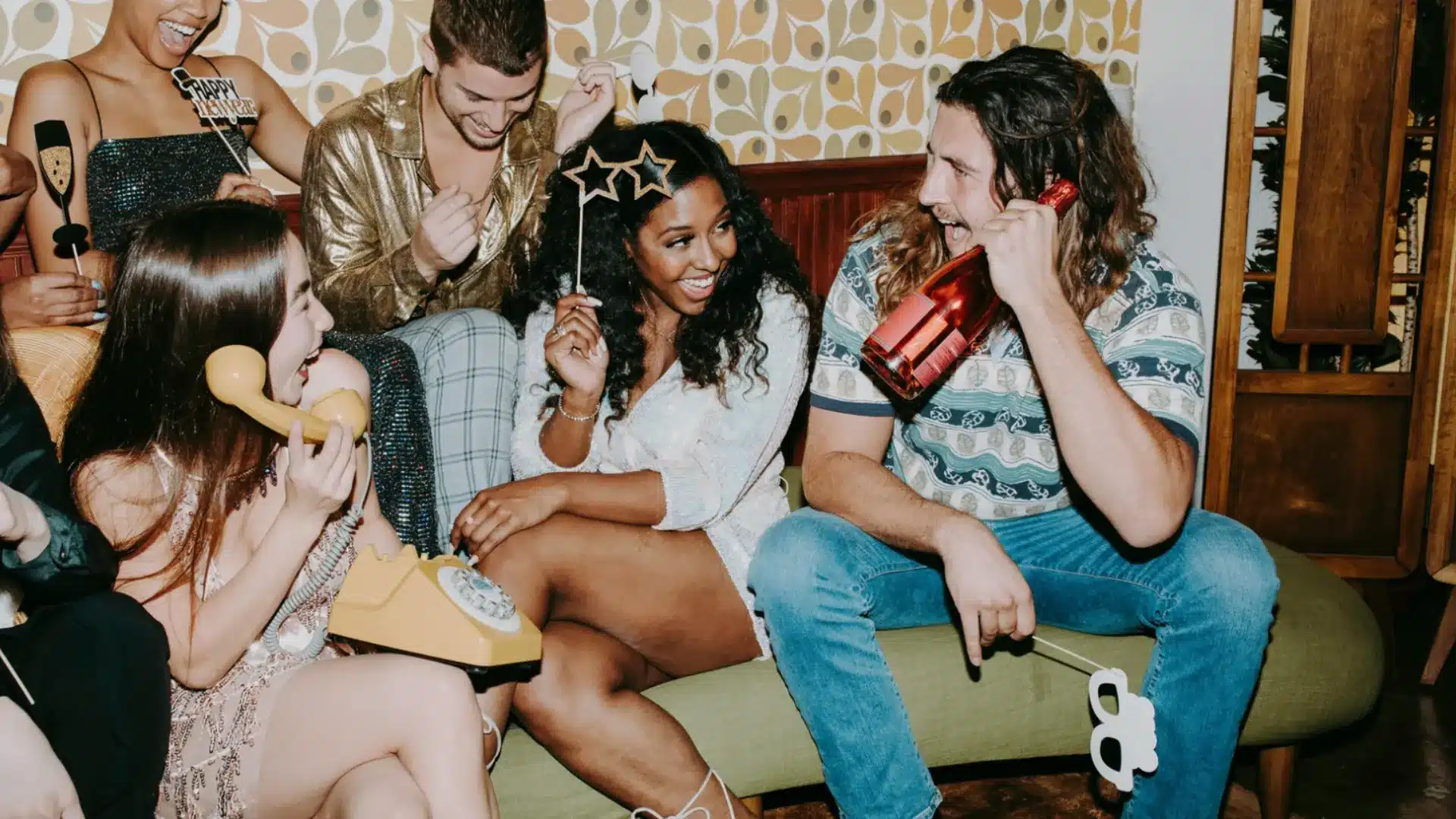 Friends celebrating at party with props, drinks, and retro decor while laughing together on a couch