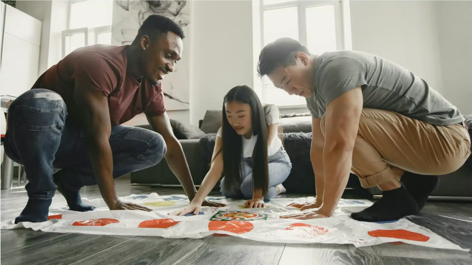 Friends playing Twister game together at home, laughing and reaching for colored spots on mat indoors