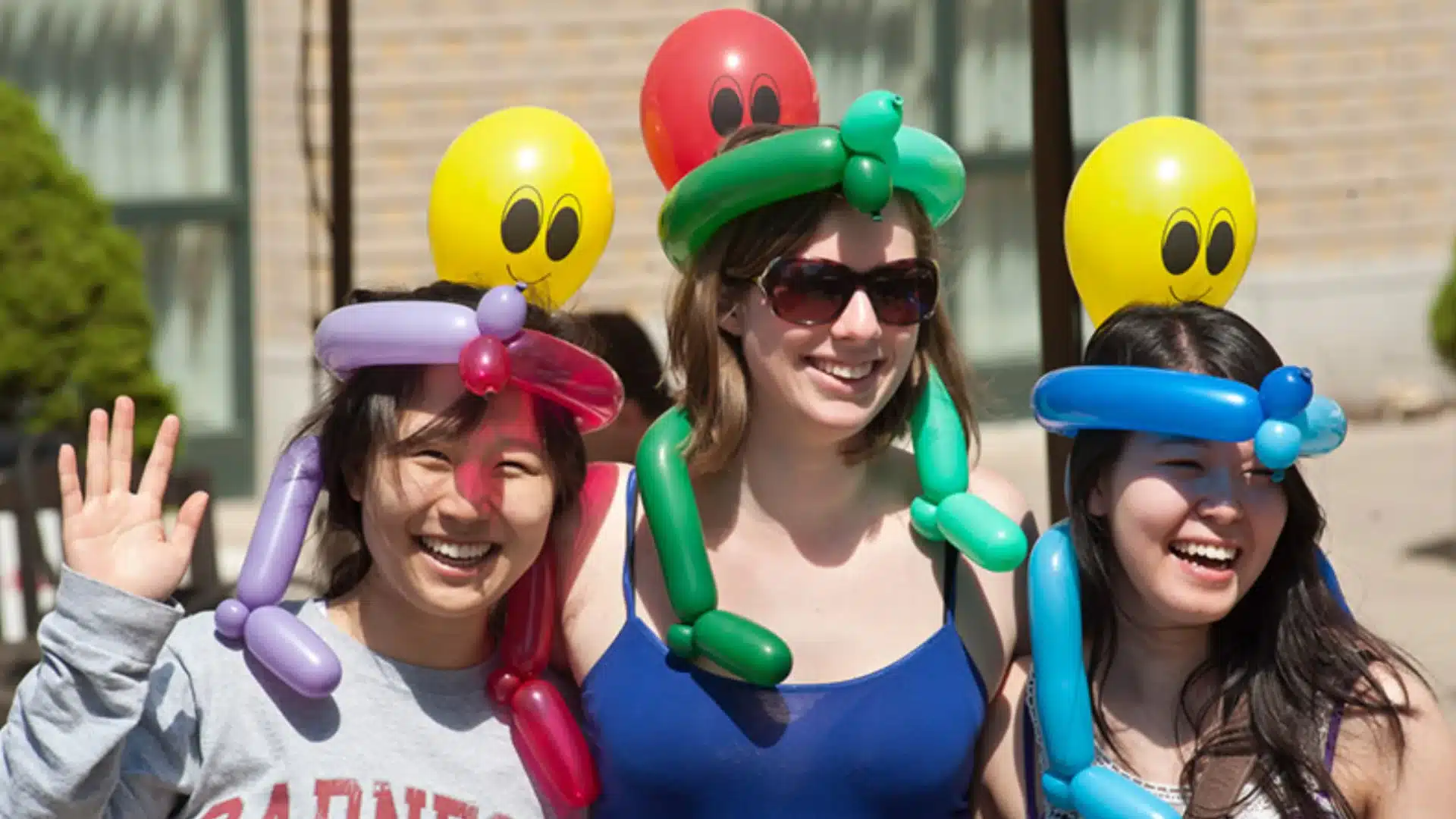 Friends smiling outdoors wearing colorful balloon hats and holding emoji balloons during a lively school carnival