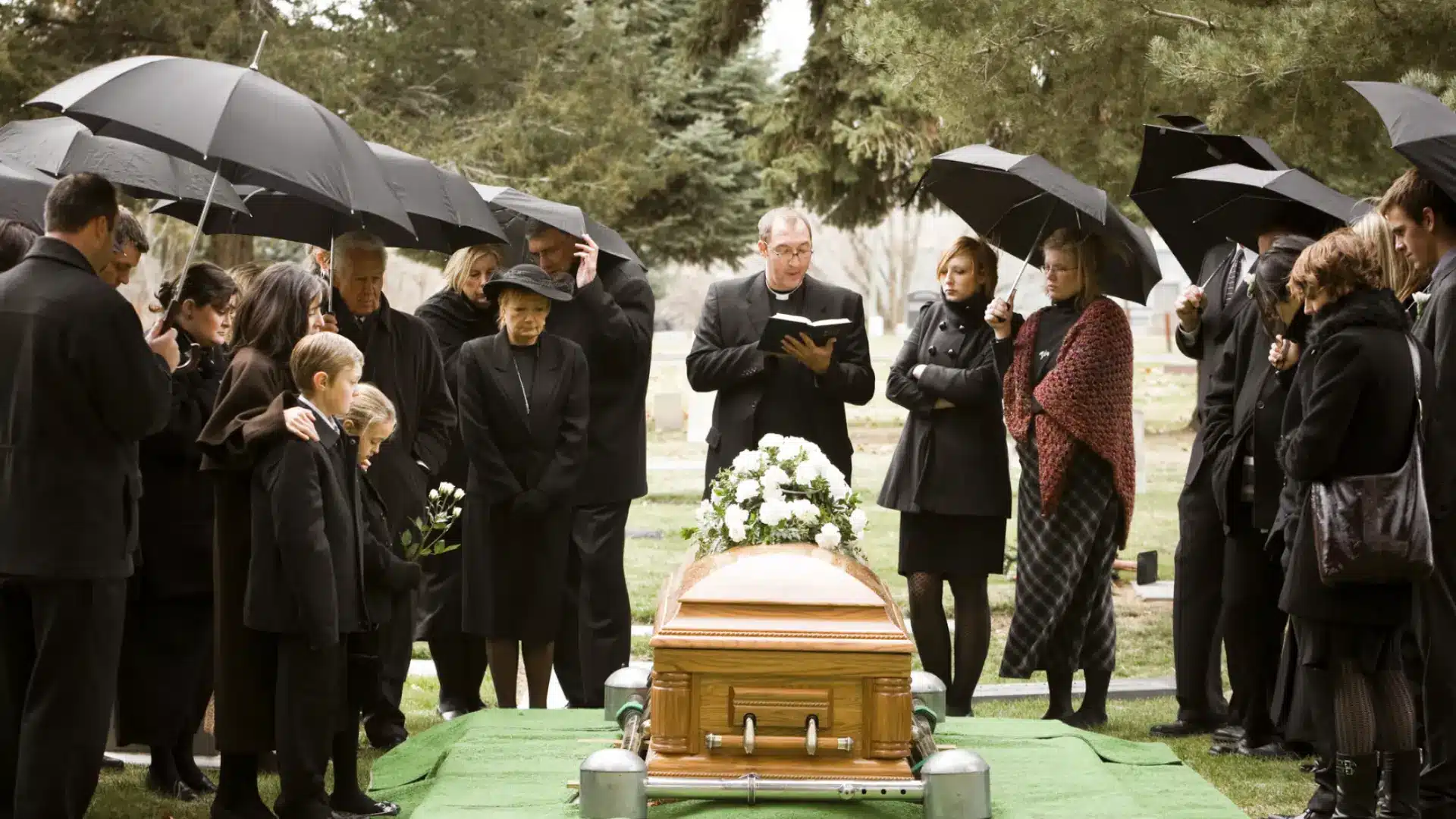 Funeral service with family gathered around casket, holding umbrellas, listening to priest during graveside ceremony