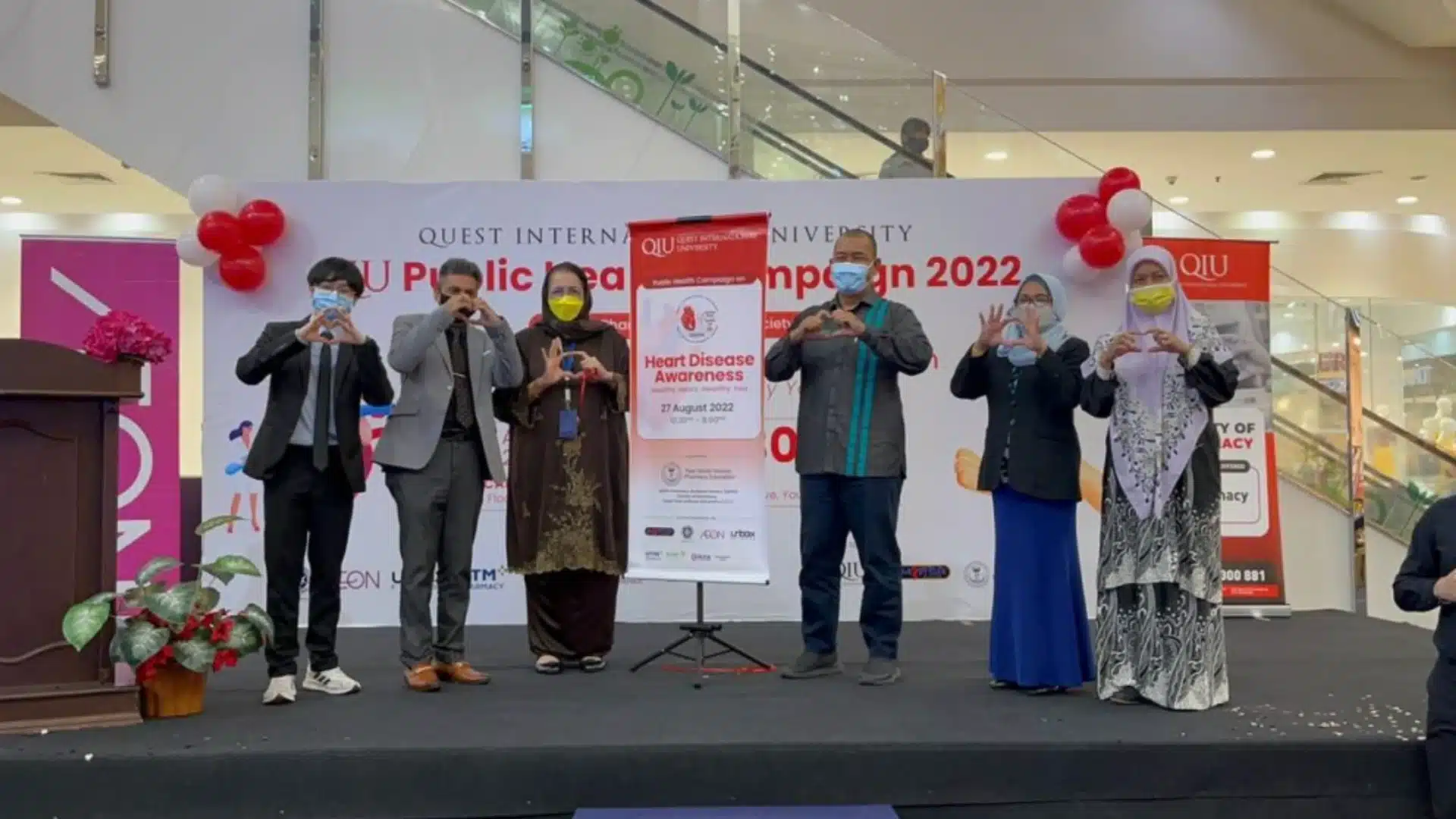 Group at public awareness campaign event holding heart signs, promoting health message on stage with banners