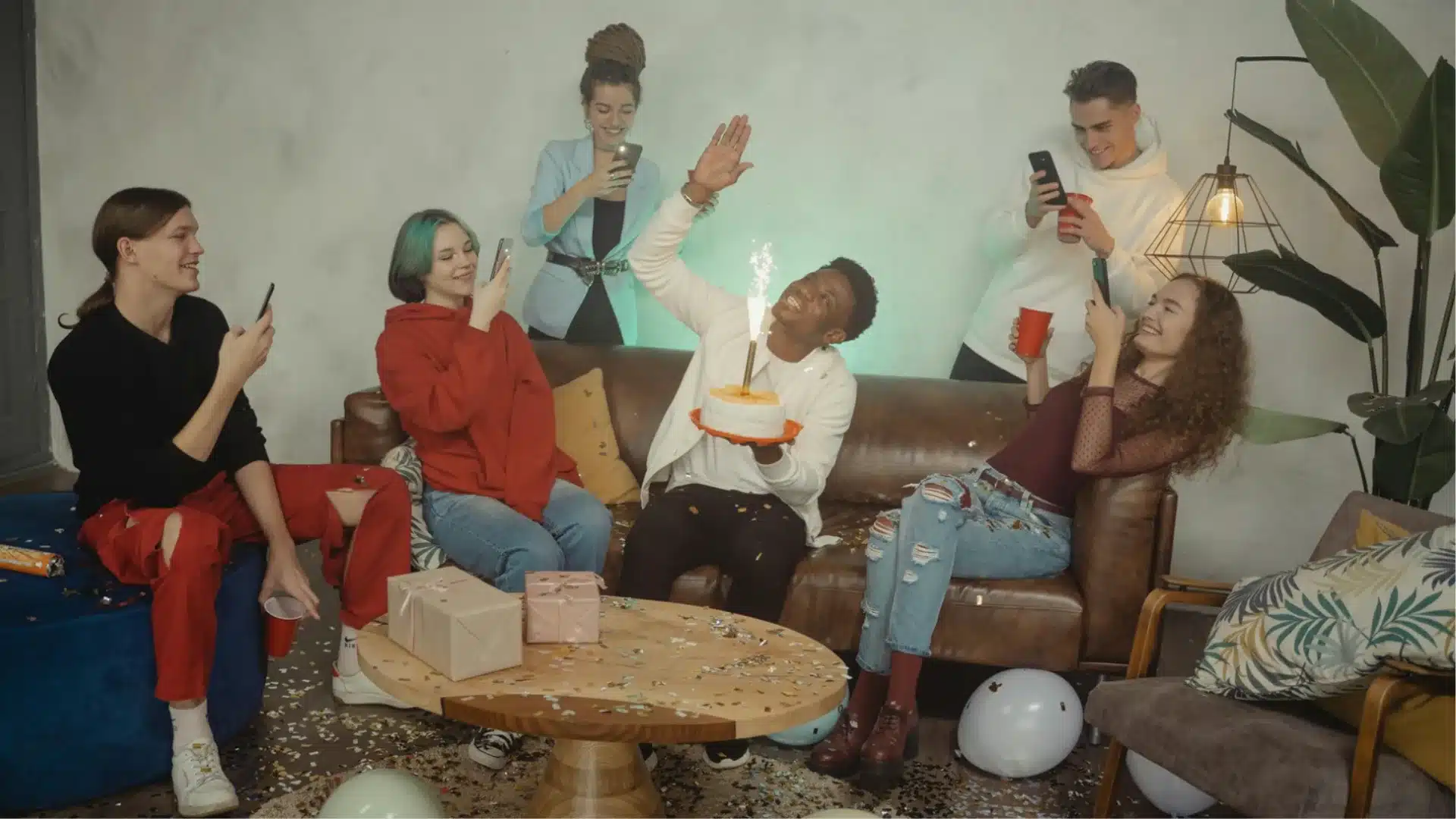 Group celebrating birthday with cake and sparkler while friends capture moment on phones indoors