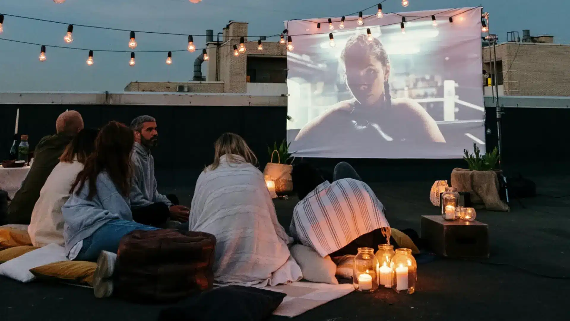 Group watching movie on rooftop at night with string lights, blankets, and cozy outdoor seating