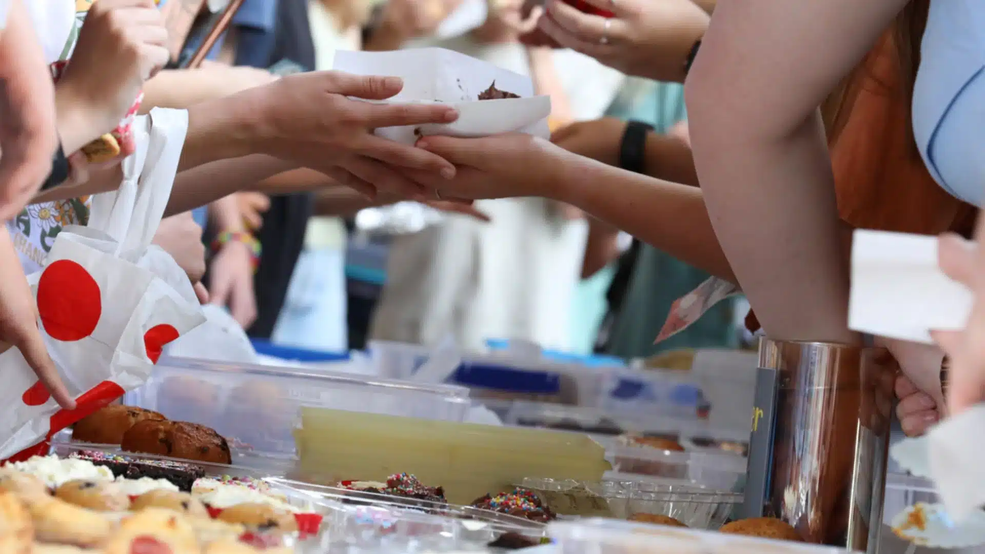 Hands exchanging baked treats at busy fundraiser table with cookies, cakes, and desserts for sale