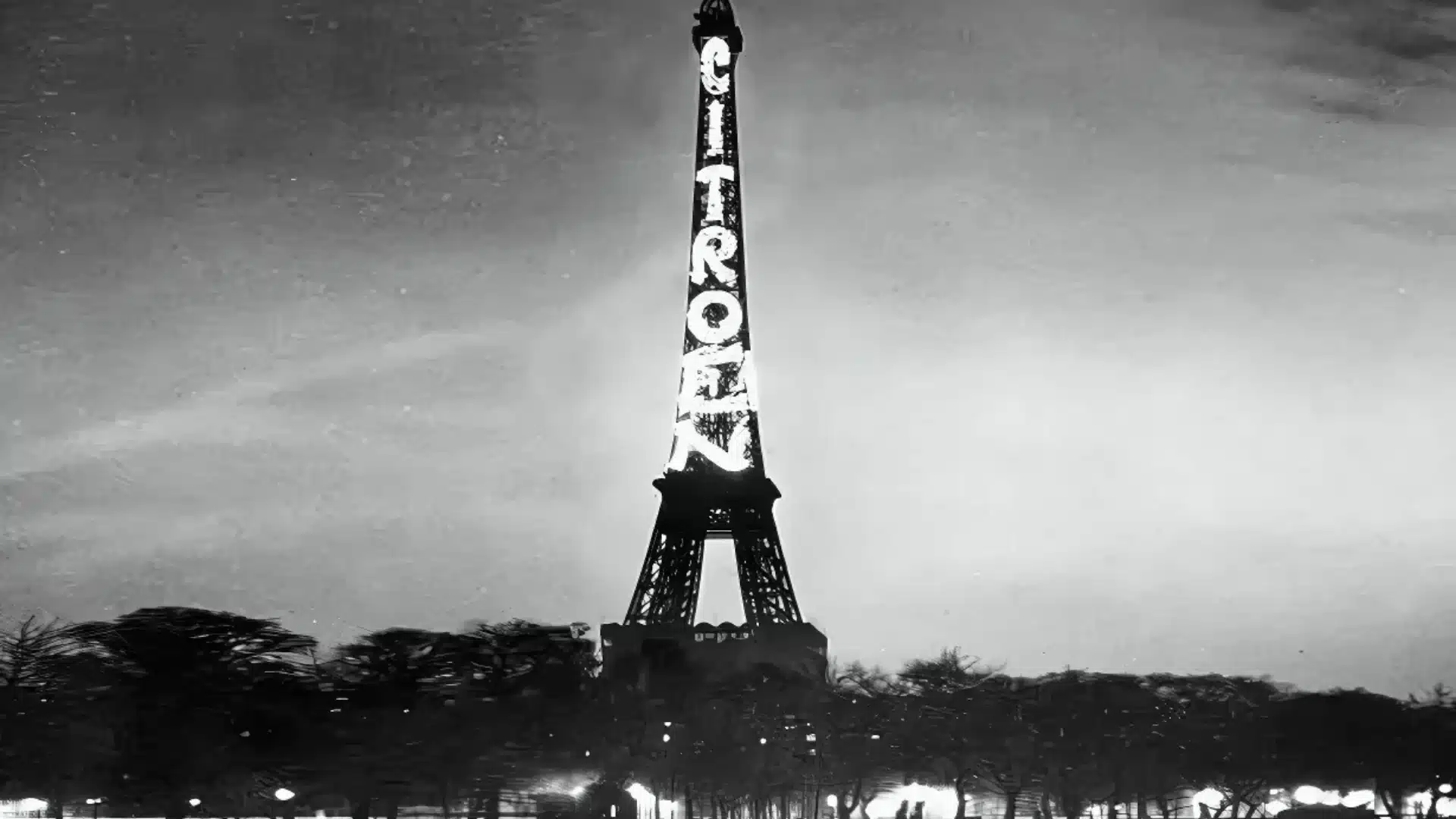 Eiffel Tower lit with large Citroën advertisement glowing at night above trees and city lights below