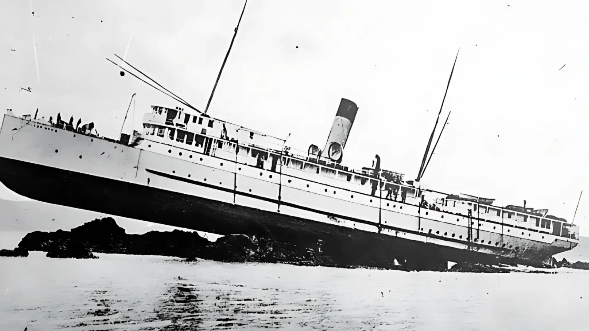 Black and white image of large steamship stranded on rocks near shore, tilted at angle during low tide