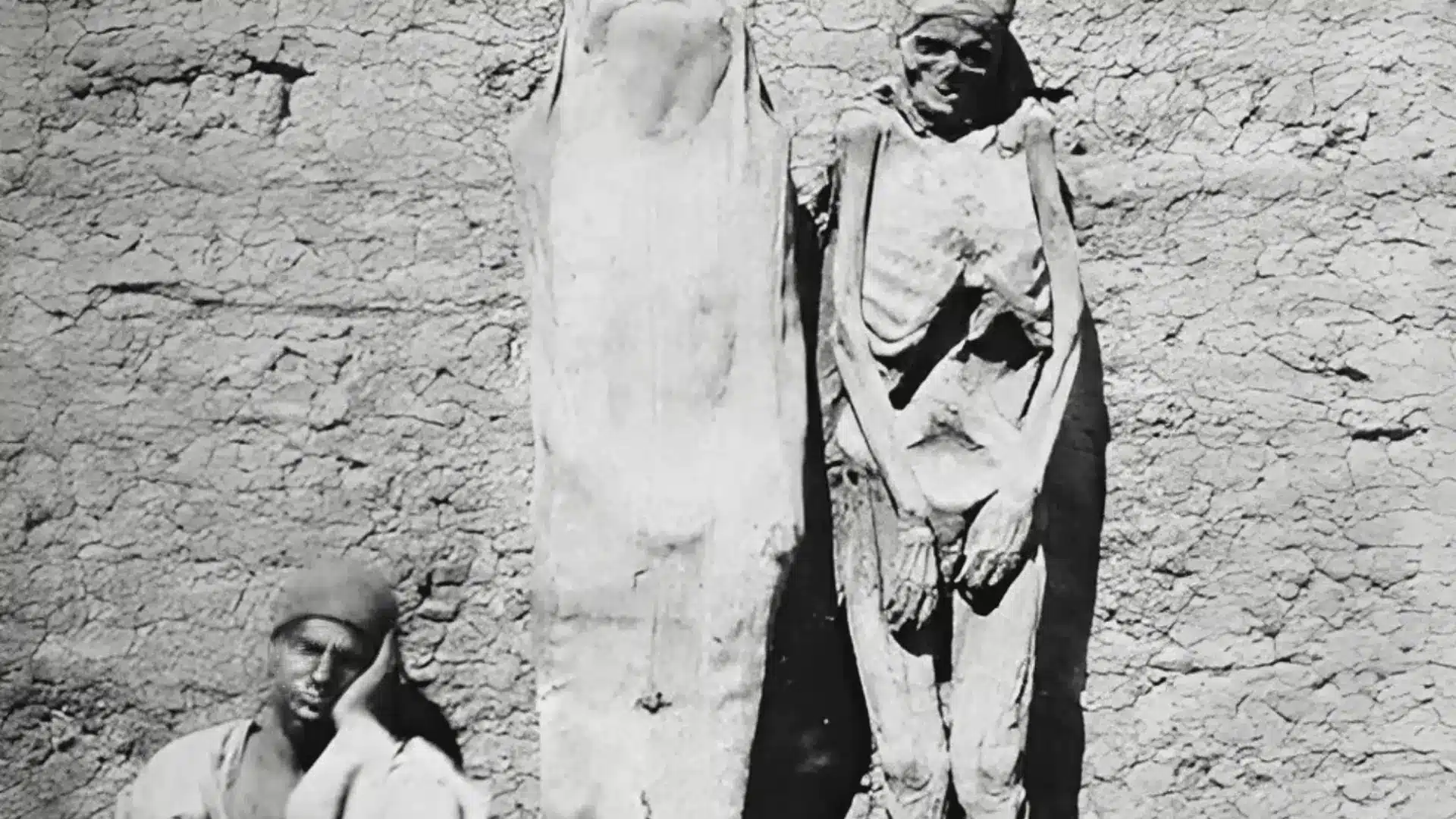 Black and white image of preserved human body displayed upright against wall with mummy nearby