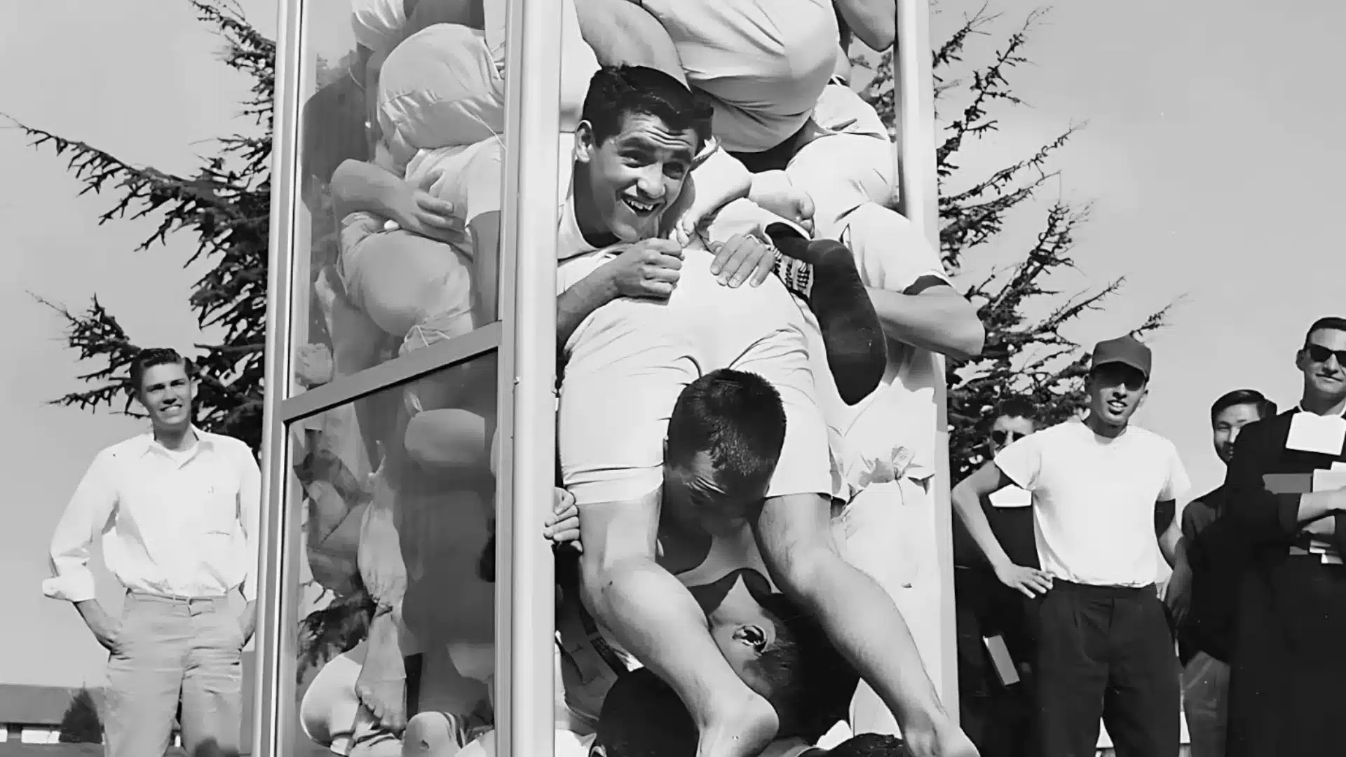 Black and white photo of men tightly packed inside phone booth during contest as crowd watches and reacts