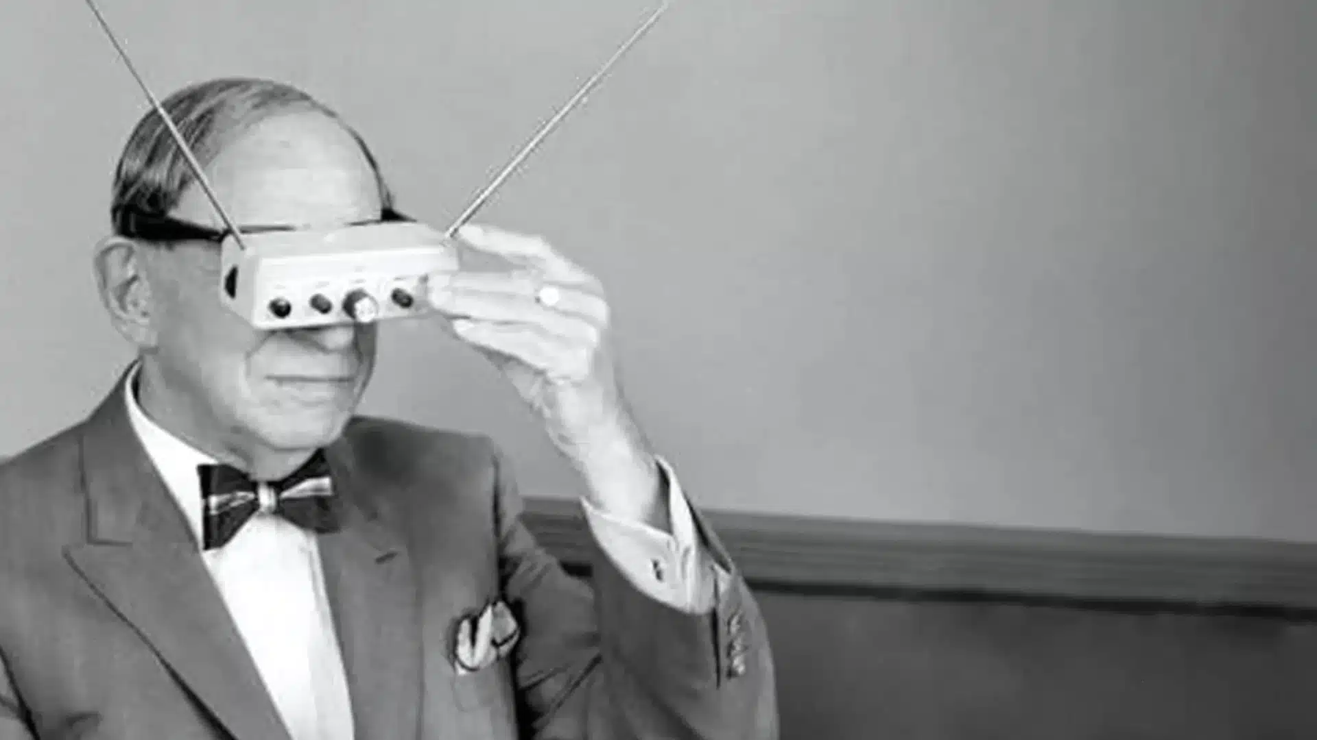 Black and white image of man wearing early television glasses device with antennas, adjusting controls