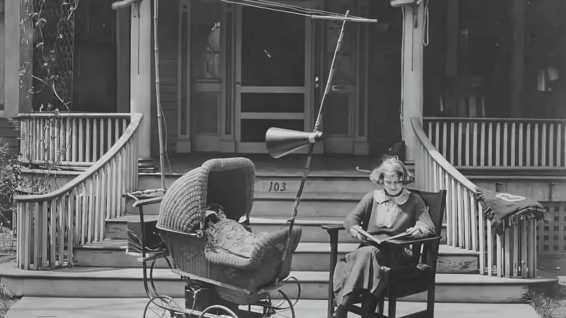 Old photo of woman reading beside baby in covered stroller outside house with mounted speaker overhead