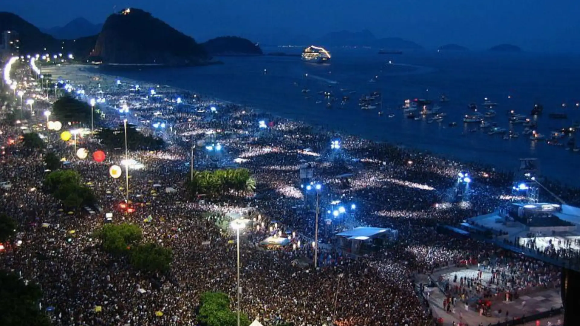 Nighttime beach concert scene at Copacabana filled with huge crowd and ocean view with boats nearby