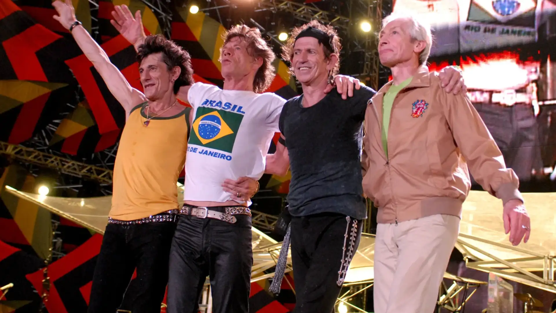 Rolling Stones band members standing together on stage waving to crowd under bright concert lights
