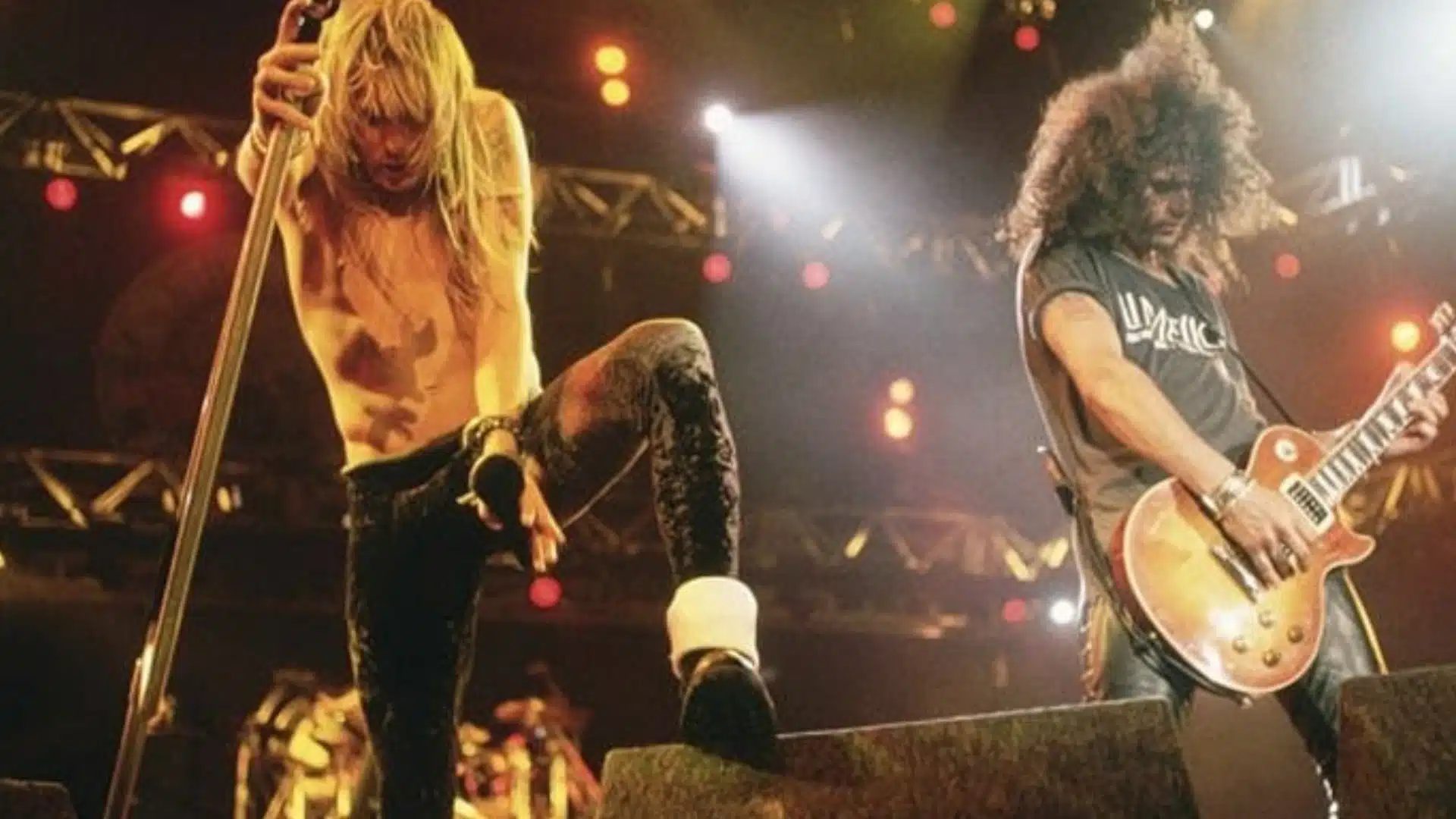 Guns N Roses performing live on stage with singer at mic stand and guitarist playing under bright lights