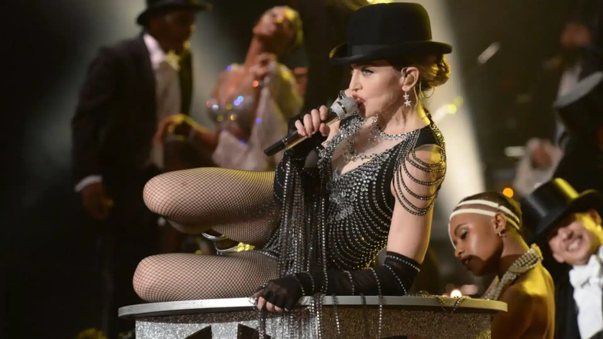 Madonna performing live in black outfit and top hat singing into microphone with dancers on stage behind her