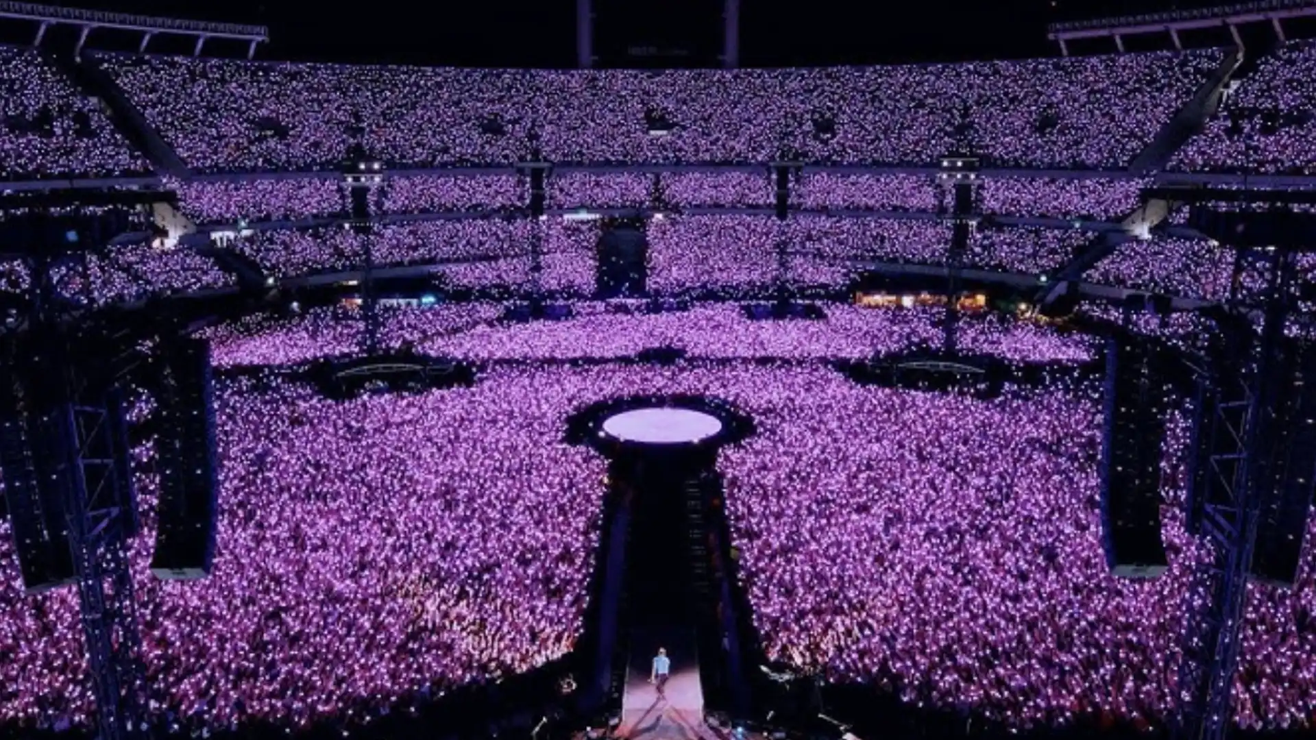 Coldplay stadium concert with crowd holding glowing lights creating sea of purple across entire arena