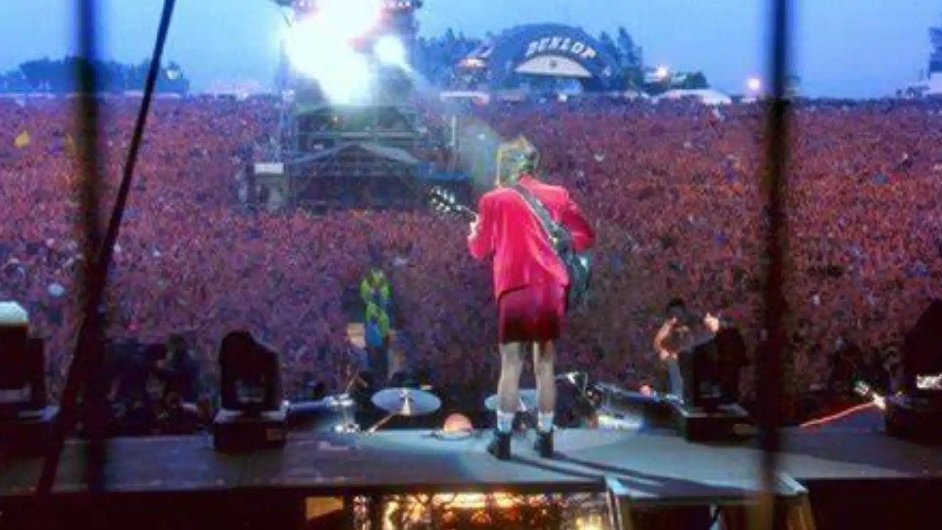AC DC performing live on stage with guitarist in red outfit facing massive crowd at outdoor festival