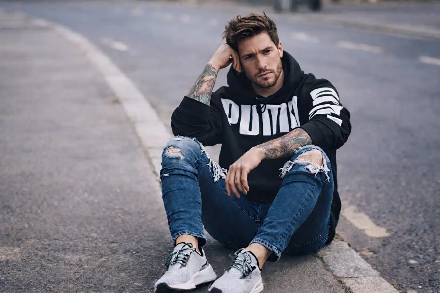 Man wearing graphic hoodie with ripped denim and sneakers sitting on street curb