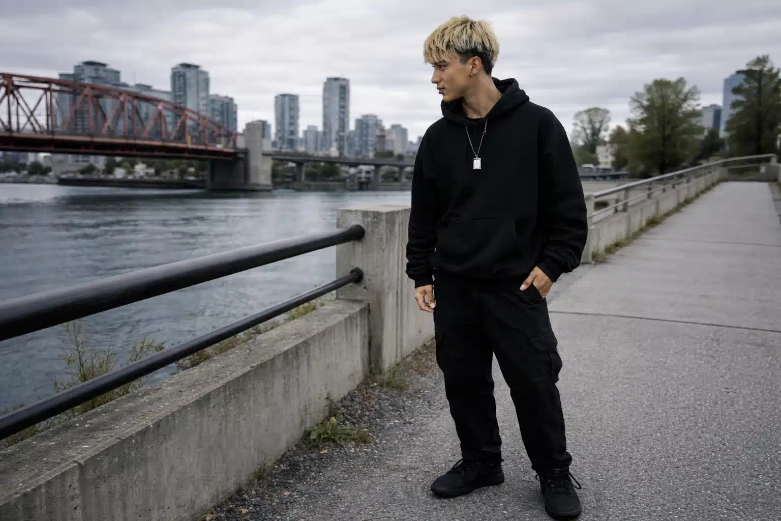 A Gen Z guy in all black outfit with bold accent necklace standing by city waterfront