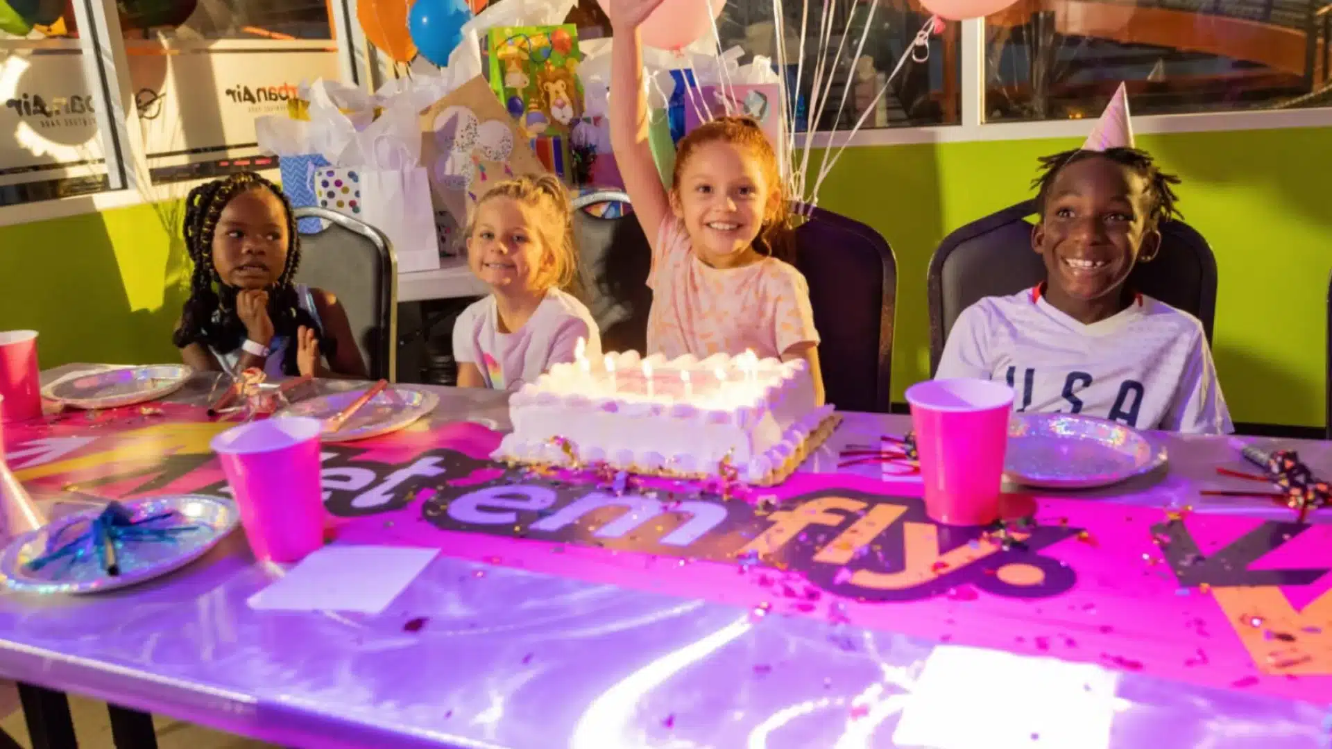 Kids celebrating birthday party with cake, balloons, and decorations at colorful indoor venue