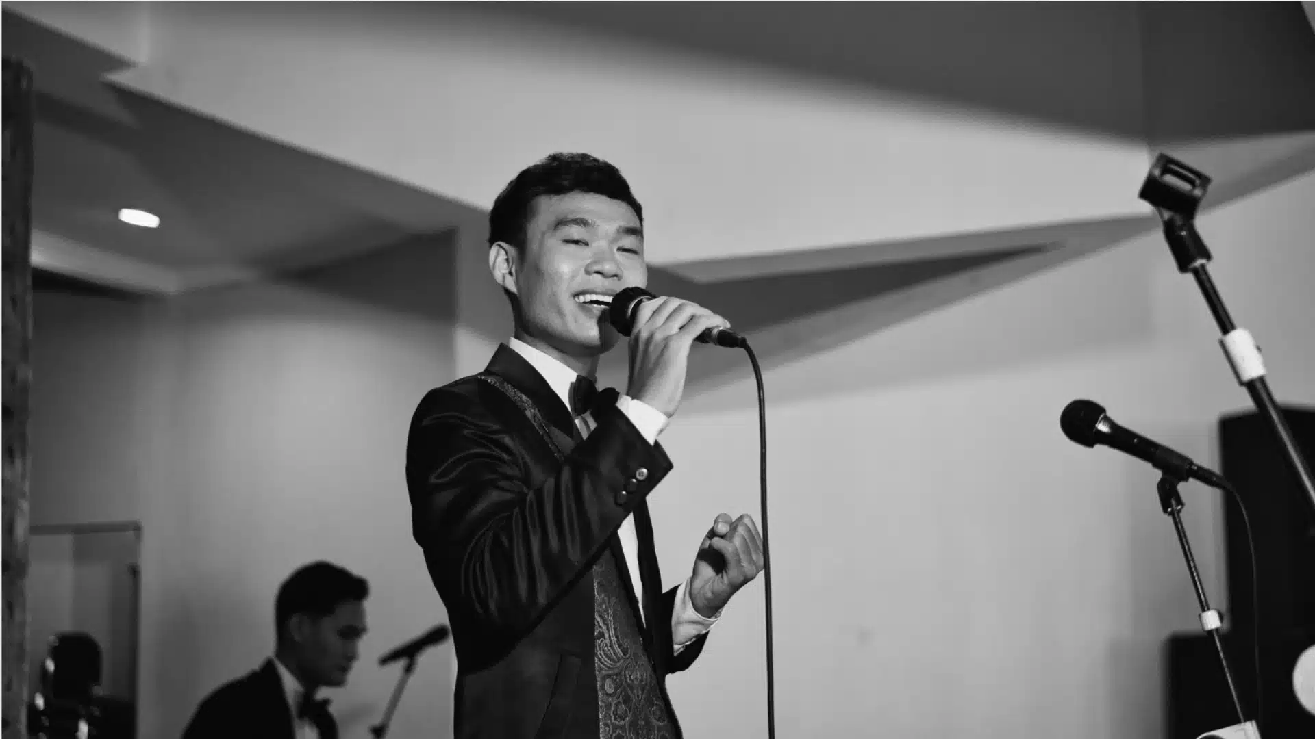 Man singing into microphone during talent show performance with band setup in indoor venue