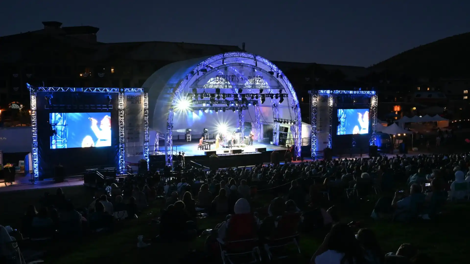 Outdoor concert at night with live band on stage, bright lights, and large seated crowd enjoying music