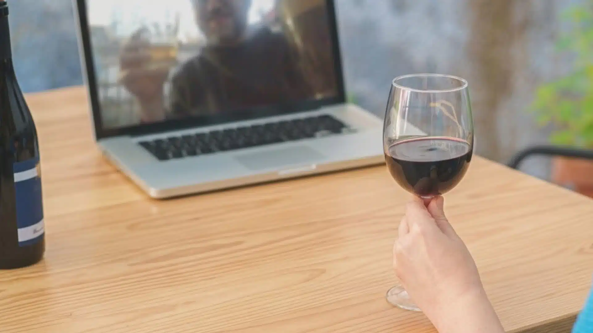 Person holding wine glass during video call on laptop, enjoying relaxed virtual hangout at home