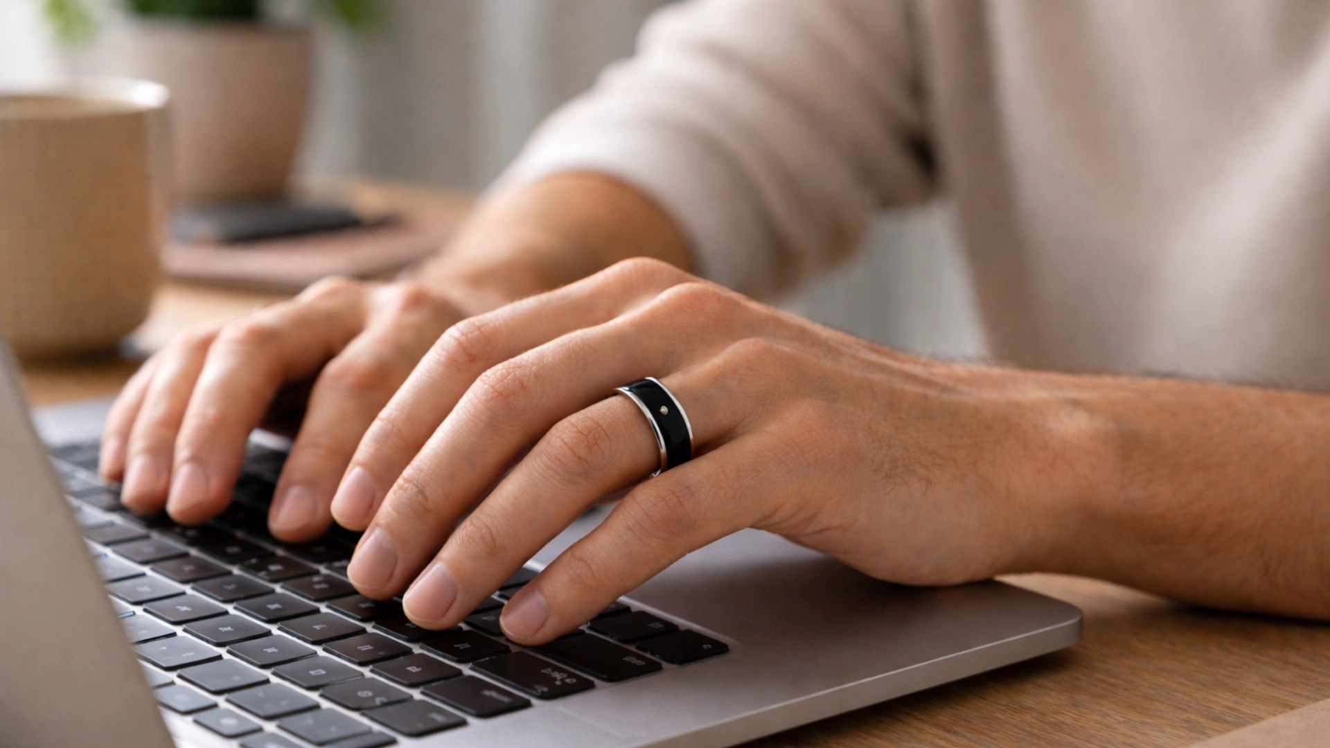 Person typing on a laptop with one hand wearing a sleek smart ring, warm desk lighting, close-up focus on fingers and keyboard