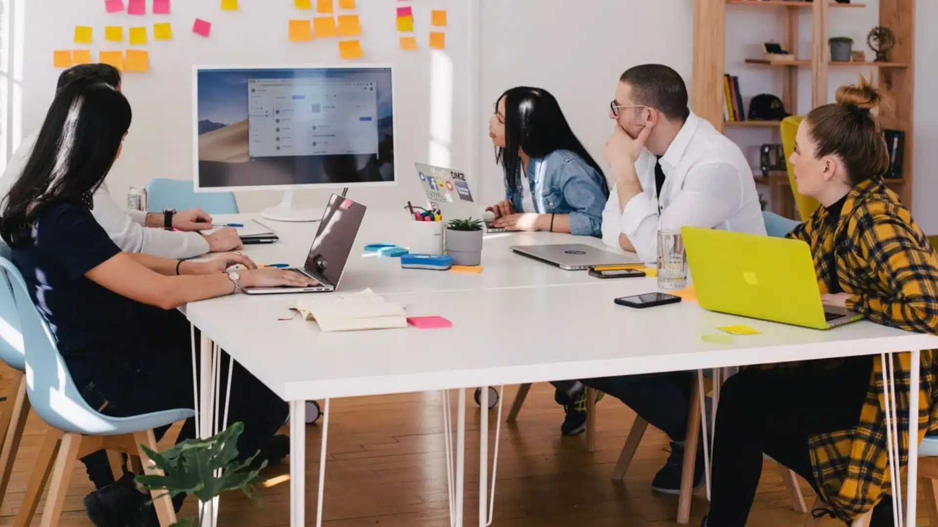 Team collaborating in office meeting around desk with laptops, notes, and presentation on screen