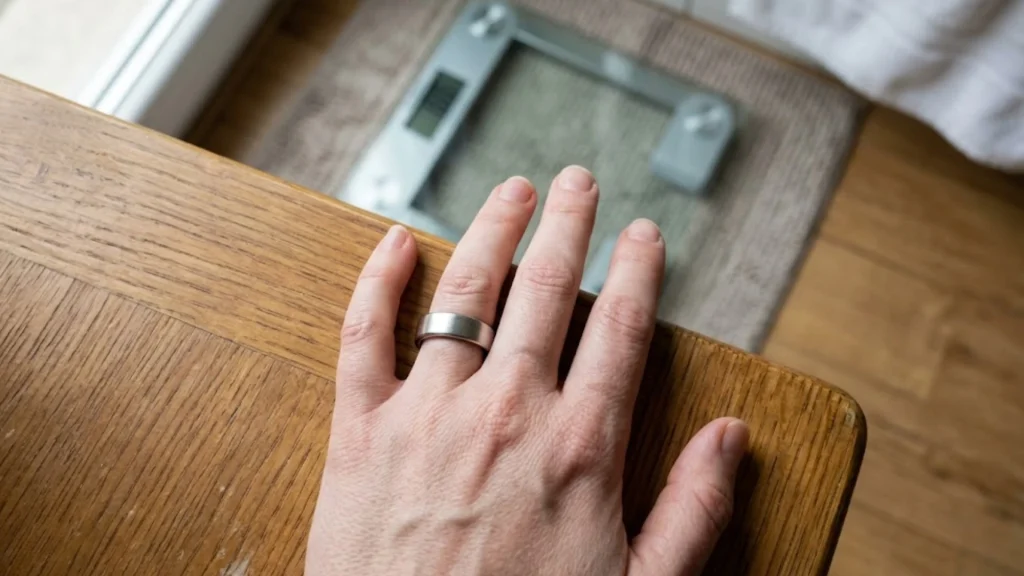 Top-down view a hand on a table with a loose smart ring and a scale, showing weight loss progress.