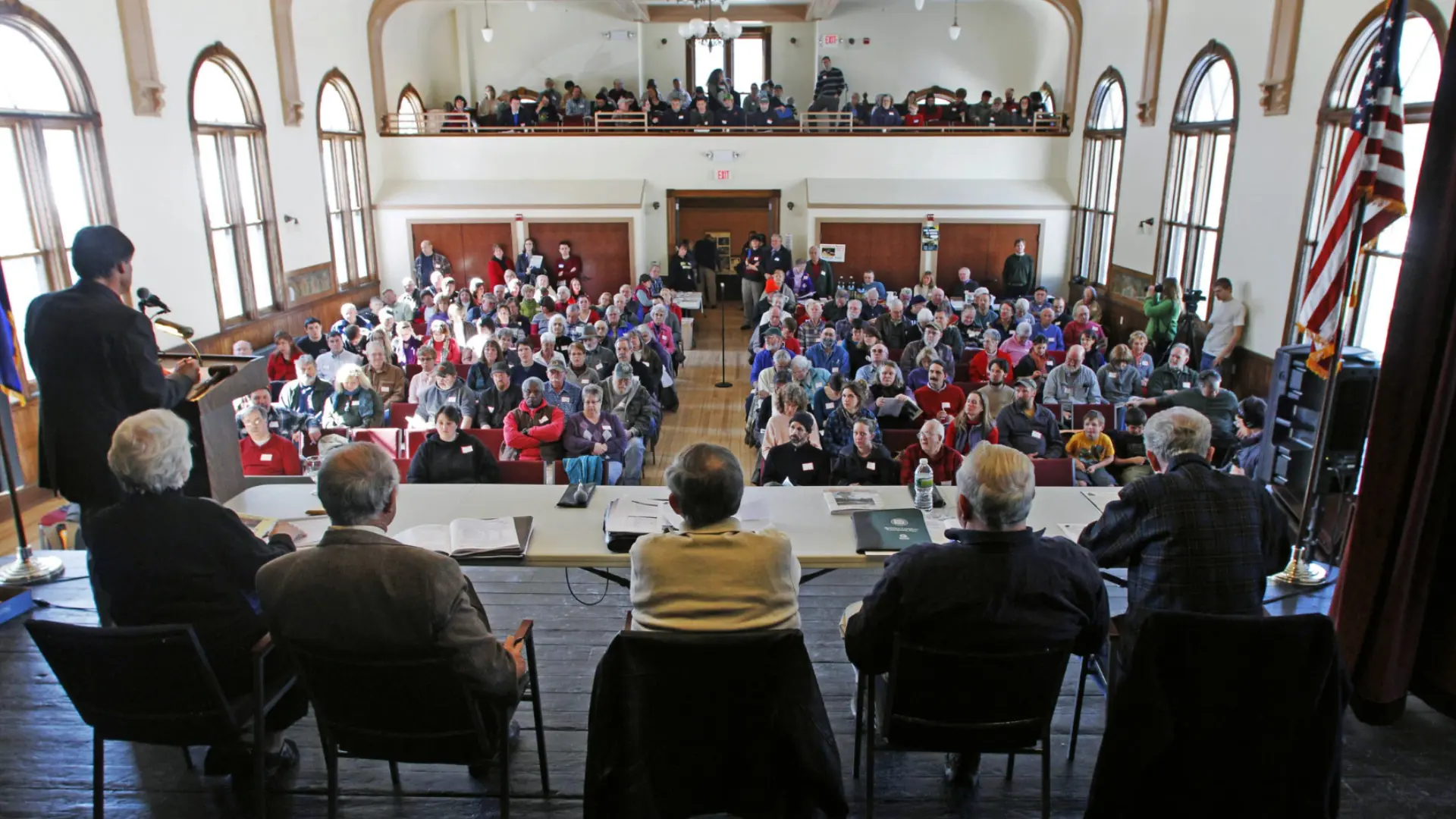 Town hall meeting with speaker at podium addressing large audience seated in community hall with balcony