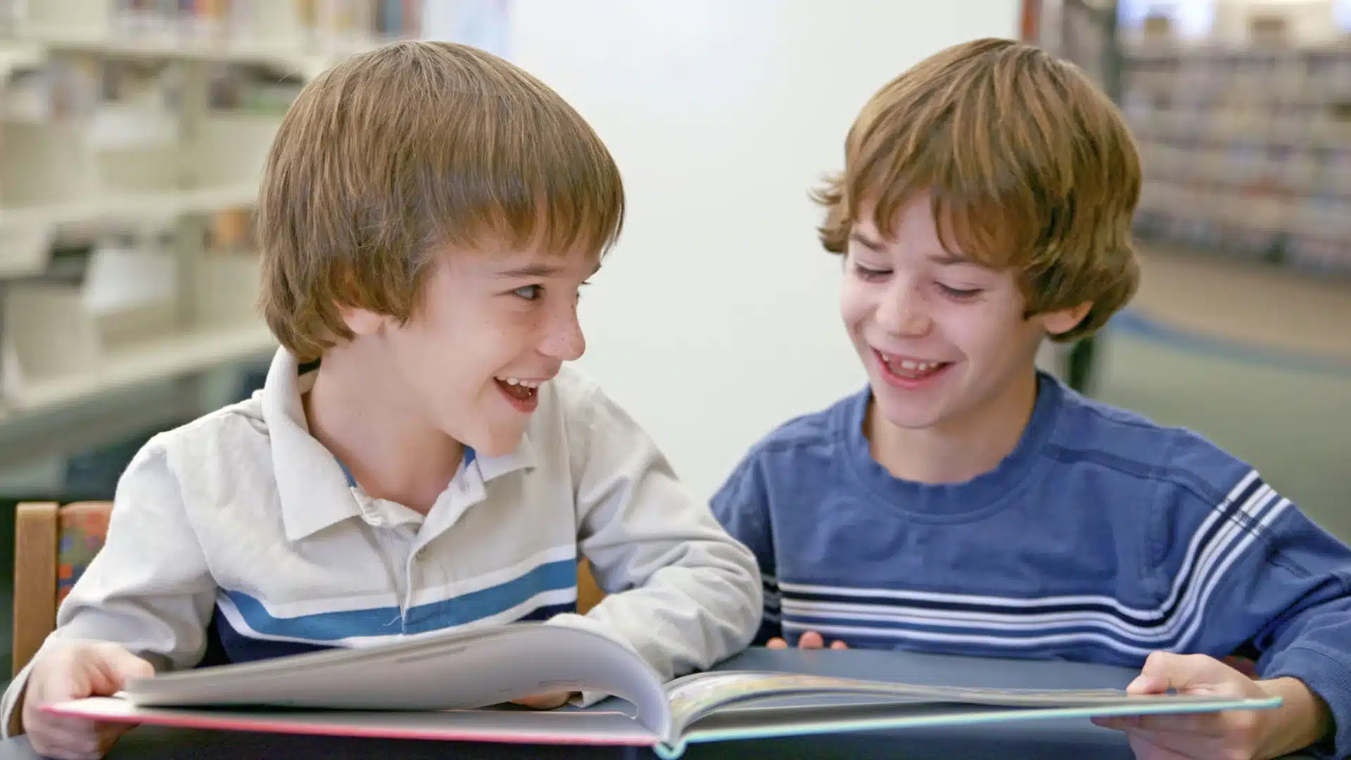Two children reading book together at table, smiling during school book club or reading fundraiser