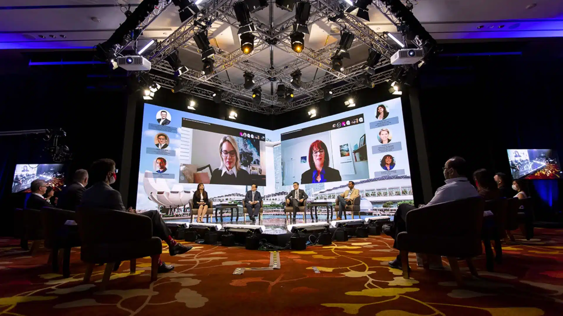 Virtual conference with panelists on stage and large screens displaying remote speakers in discussion