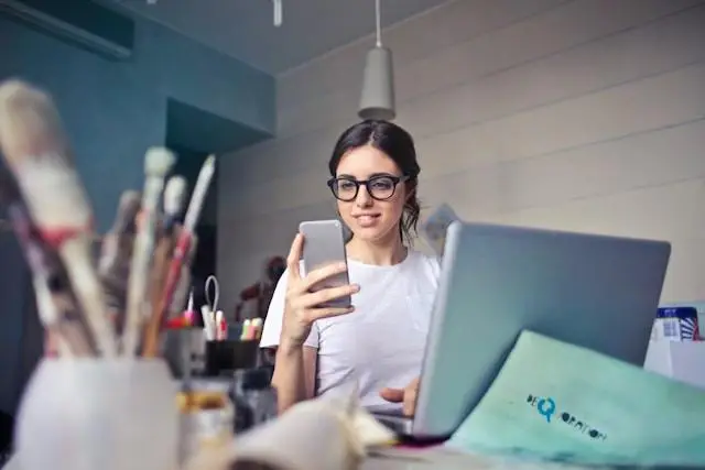 Woman using smartphone at desk with laptop and art supplies in bright workspace