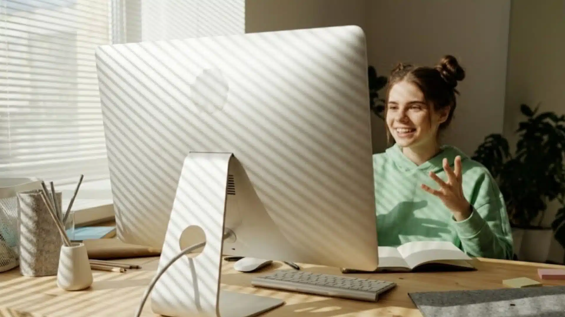 Young woman smiling and attending virtual charity event at desktop computer in sunlit room with minimal workspace