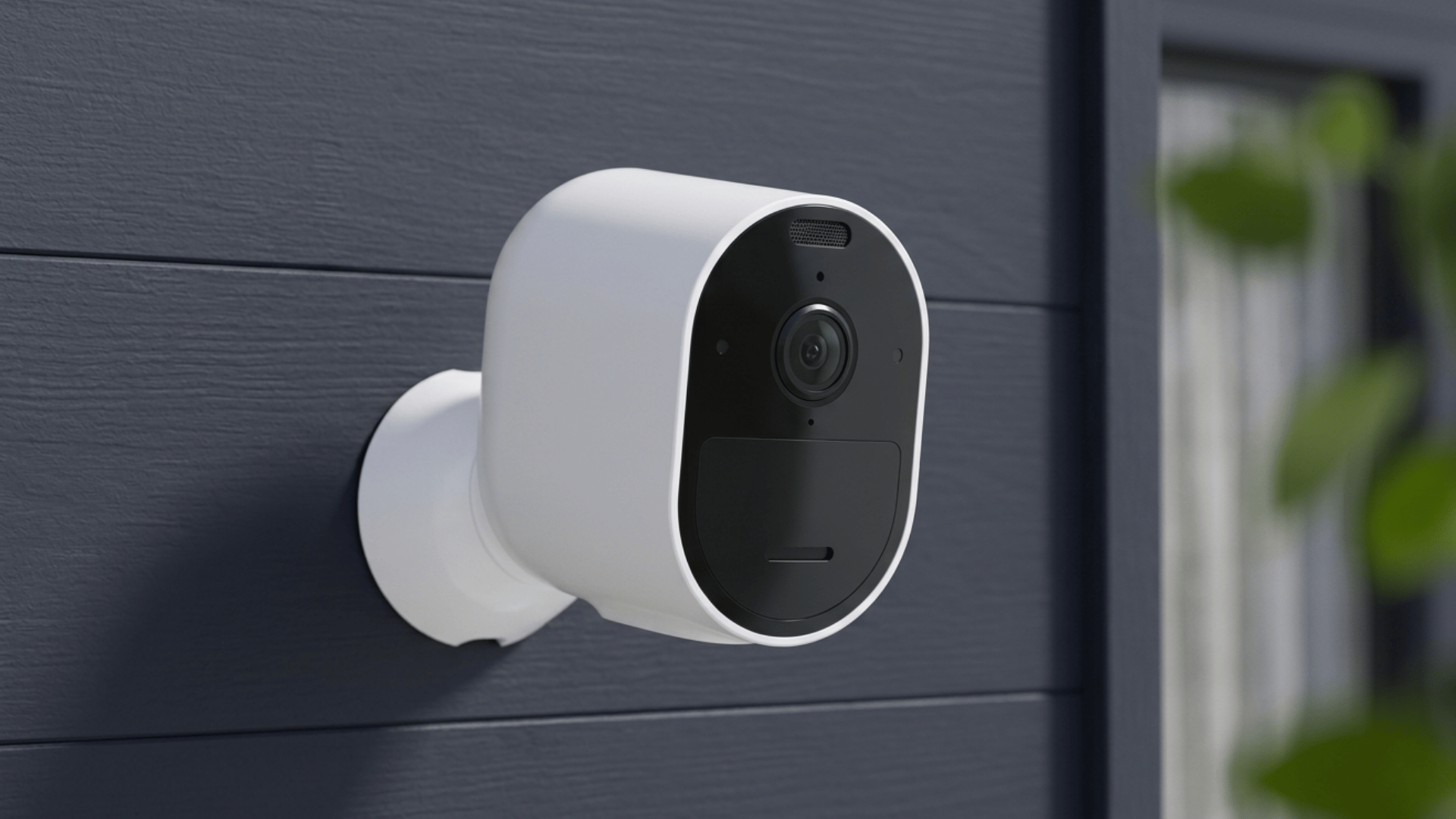 A security camera mounted on the exterior wall of a house with a blurred window and greenery in the background