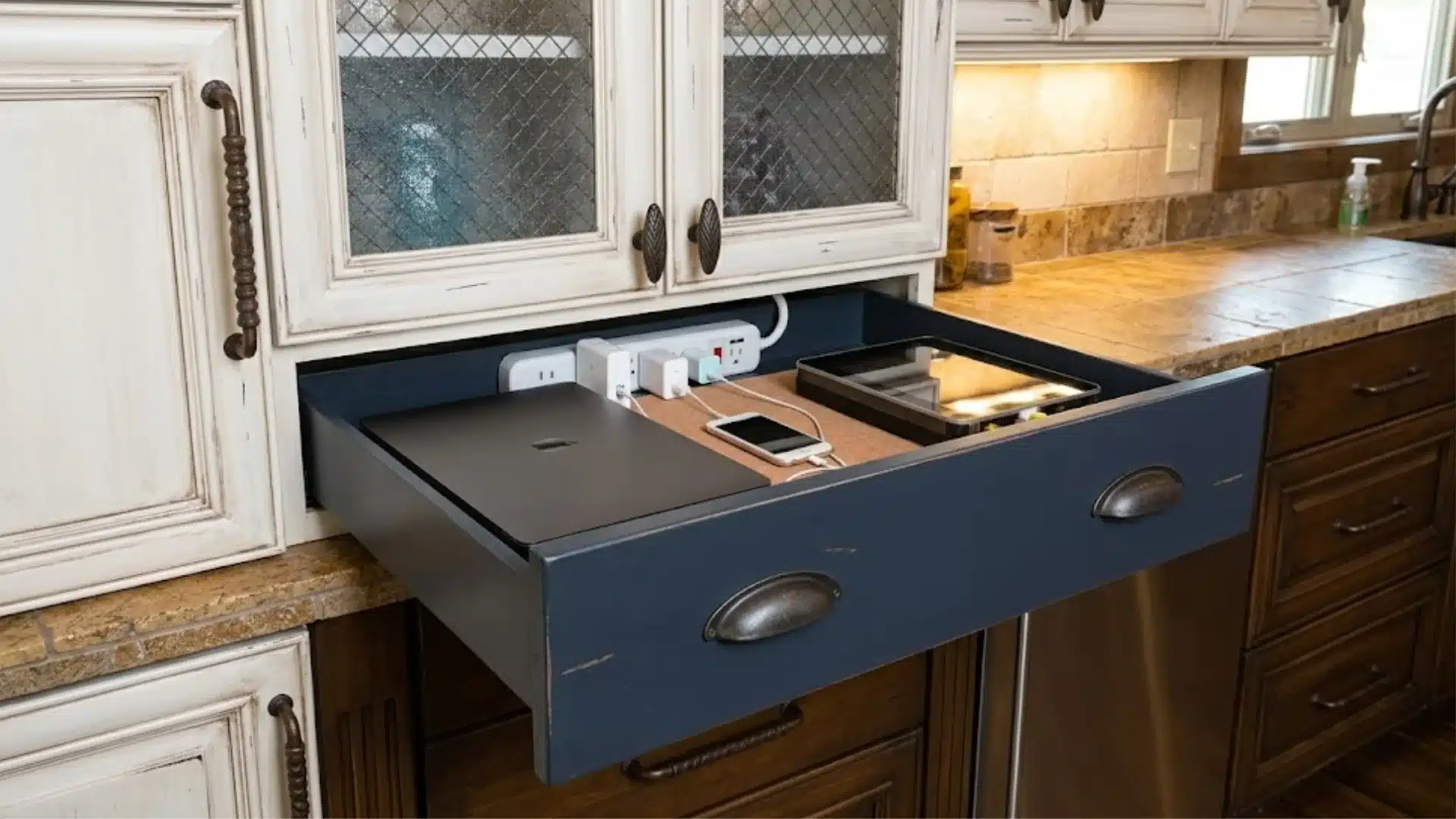 An open charging drawer in a kitchen cabinet containing a laptop, phone, and tablet plugged into a power strip