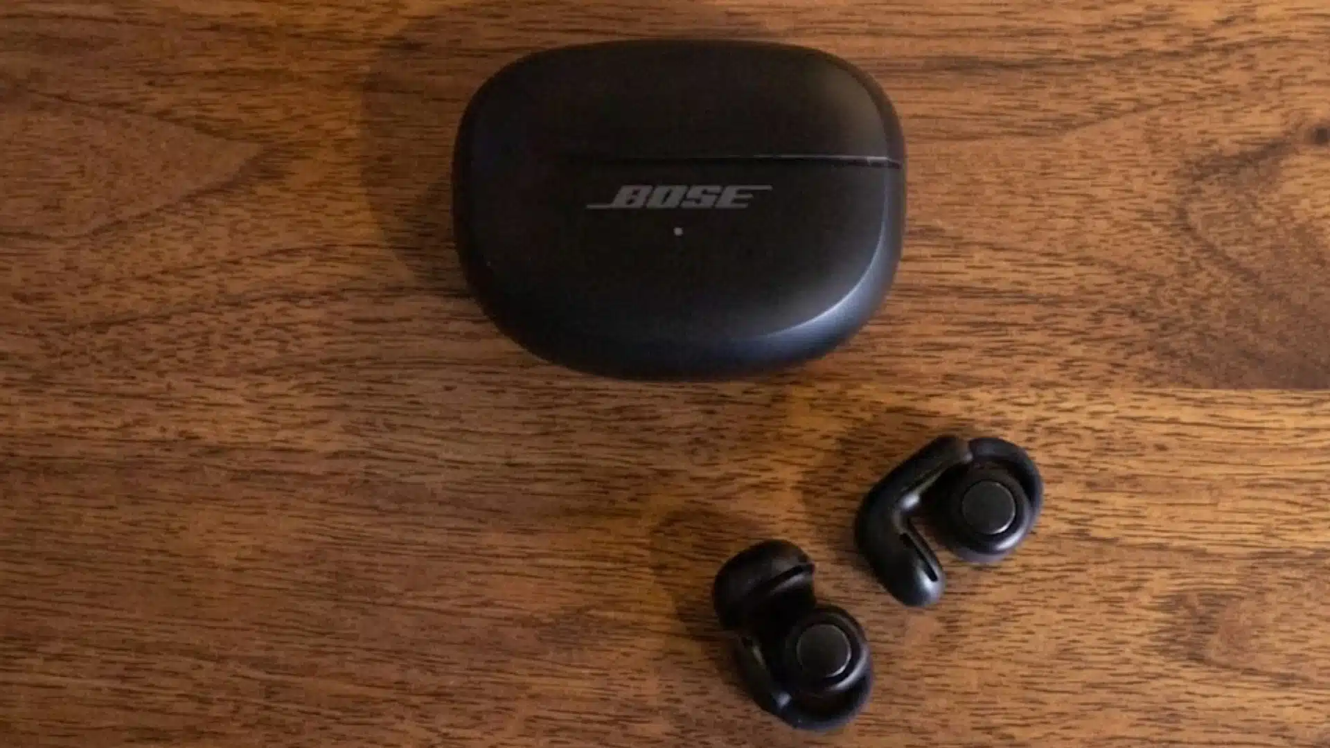 Bose Ultra Open Earbuds with their charging case displayed on a wooden surface, showing full setup