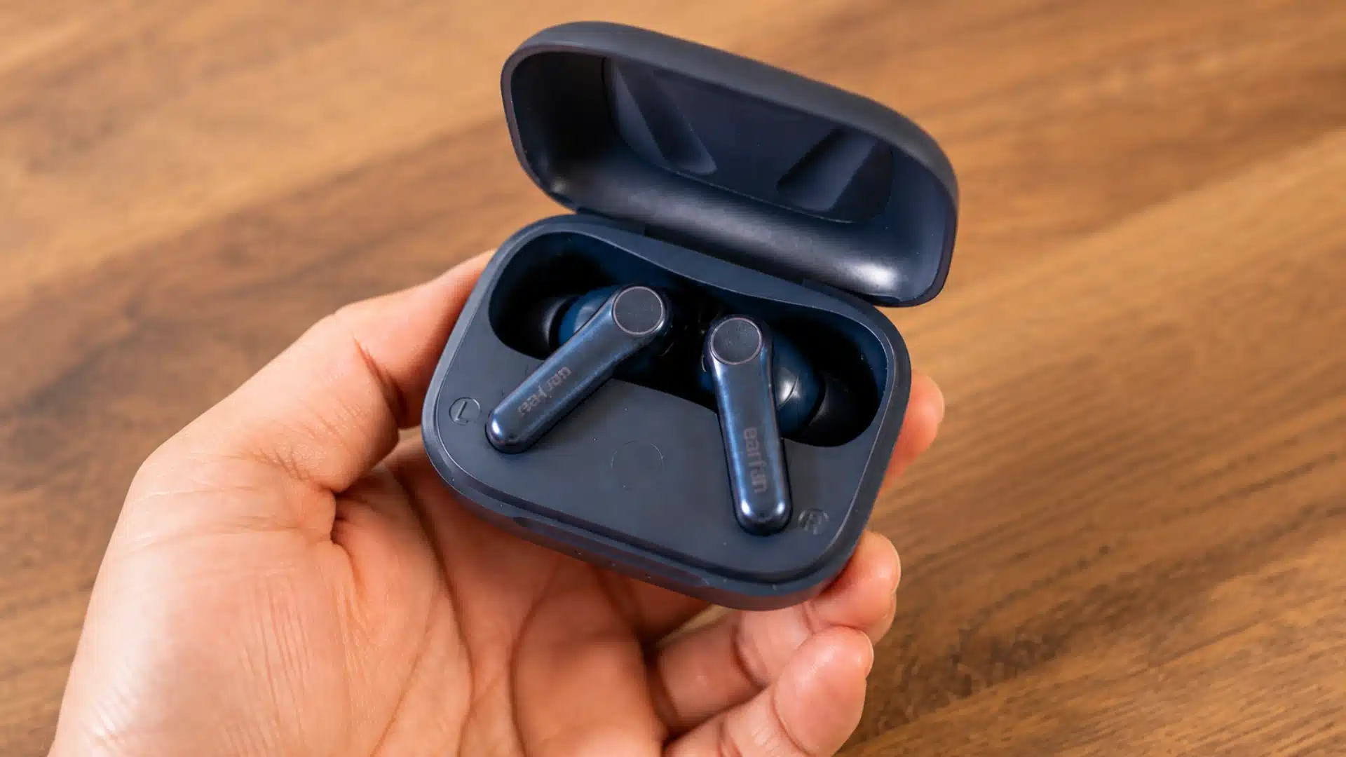 Hand holding a pair of dark wireless earbuds in an open charging case on a wooden table