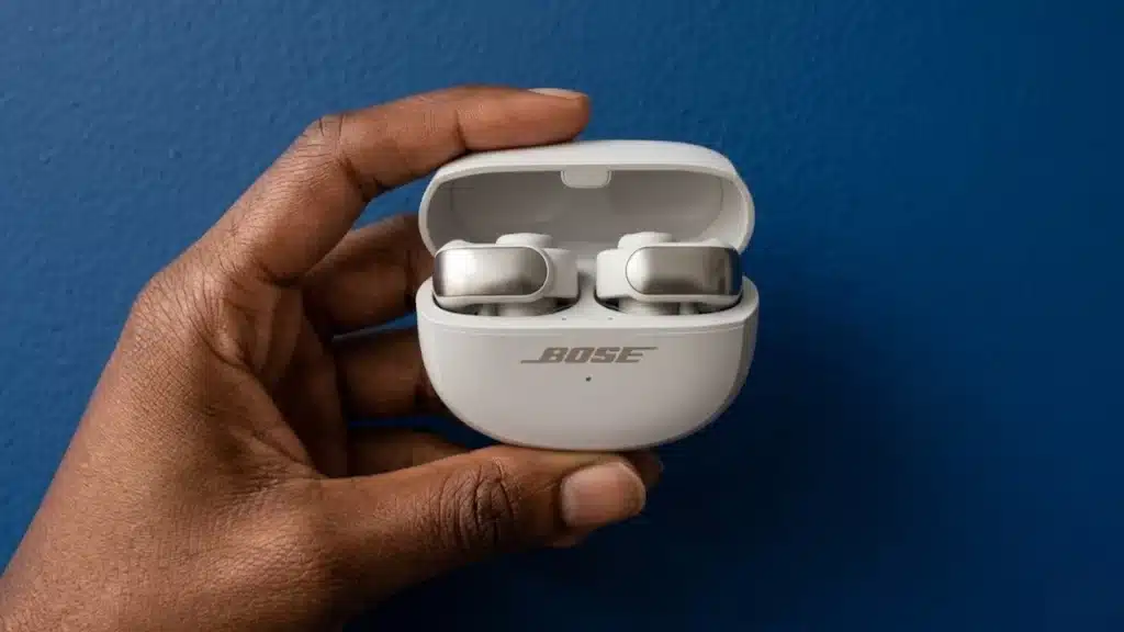 Hand holding white Bose Ultra Open Earbuds in charging case against blue background, showing earbuds clearly