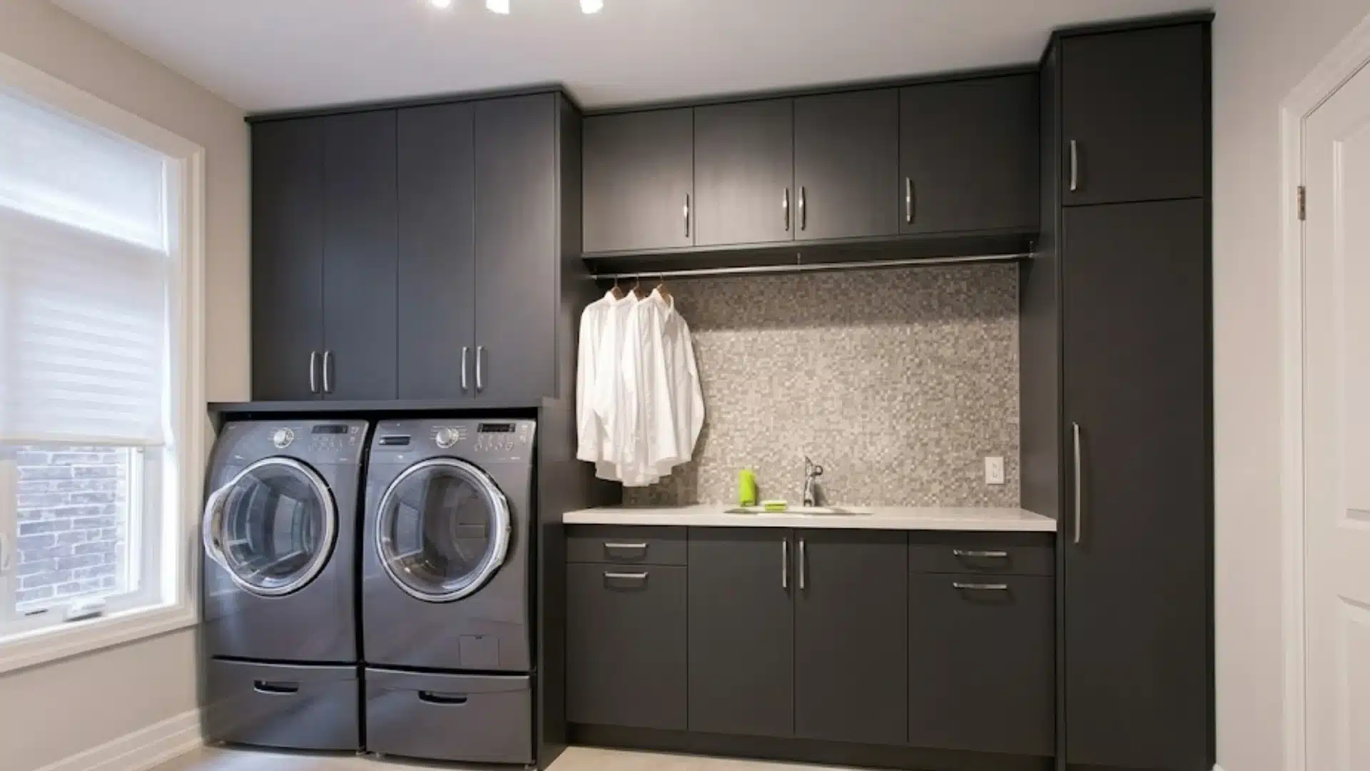 Modern laundry room with dark gray cabinets, stacked above a washer and dryer, next to a sink and countertop