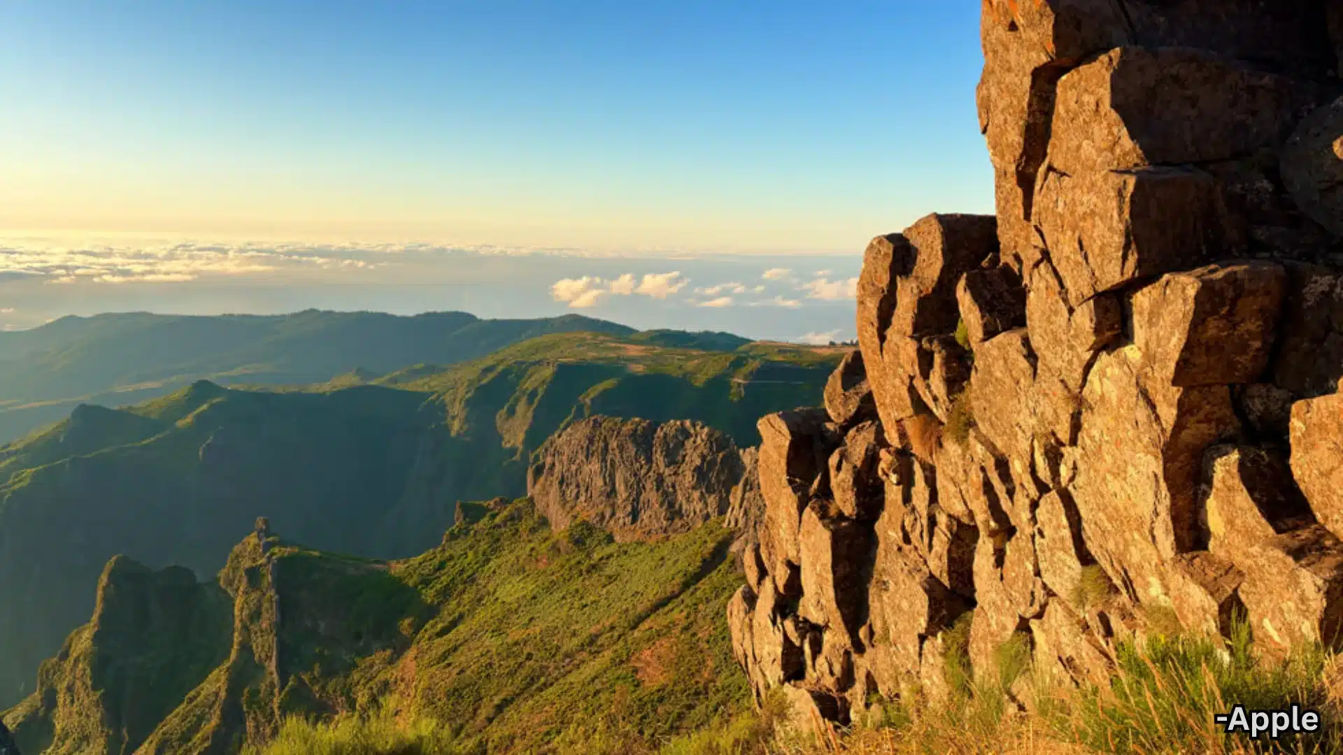 Aerial view of mountain cliffs with rocky formations, green valleys, and clouds under clear blue sky during golden hour