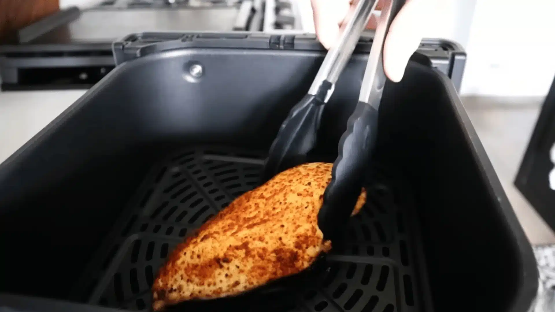 Air fryer basket holding seasoned chicken breast with silicone tongs during cooking