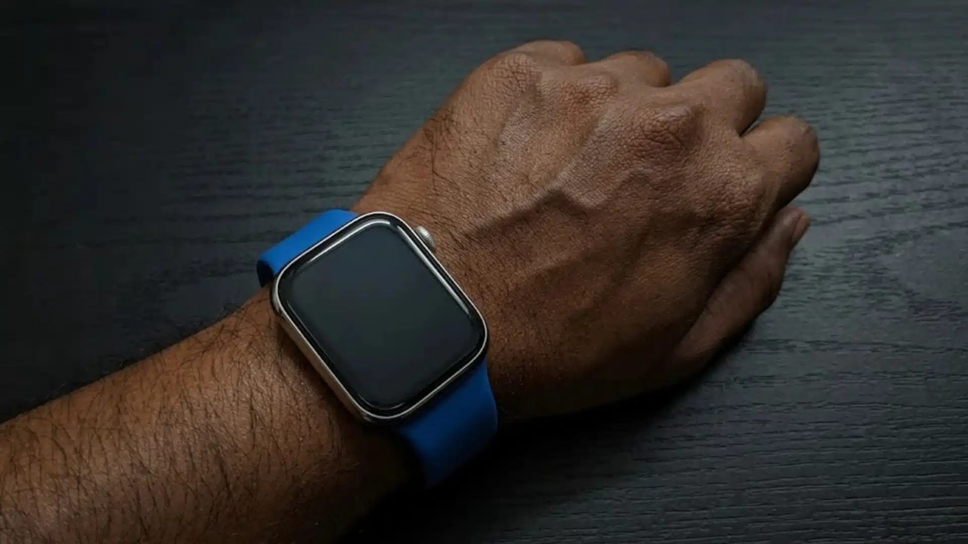 Apple Watch worn on wrist with blue band, showing large rectangular display on a person’s hand on dark surface