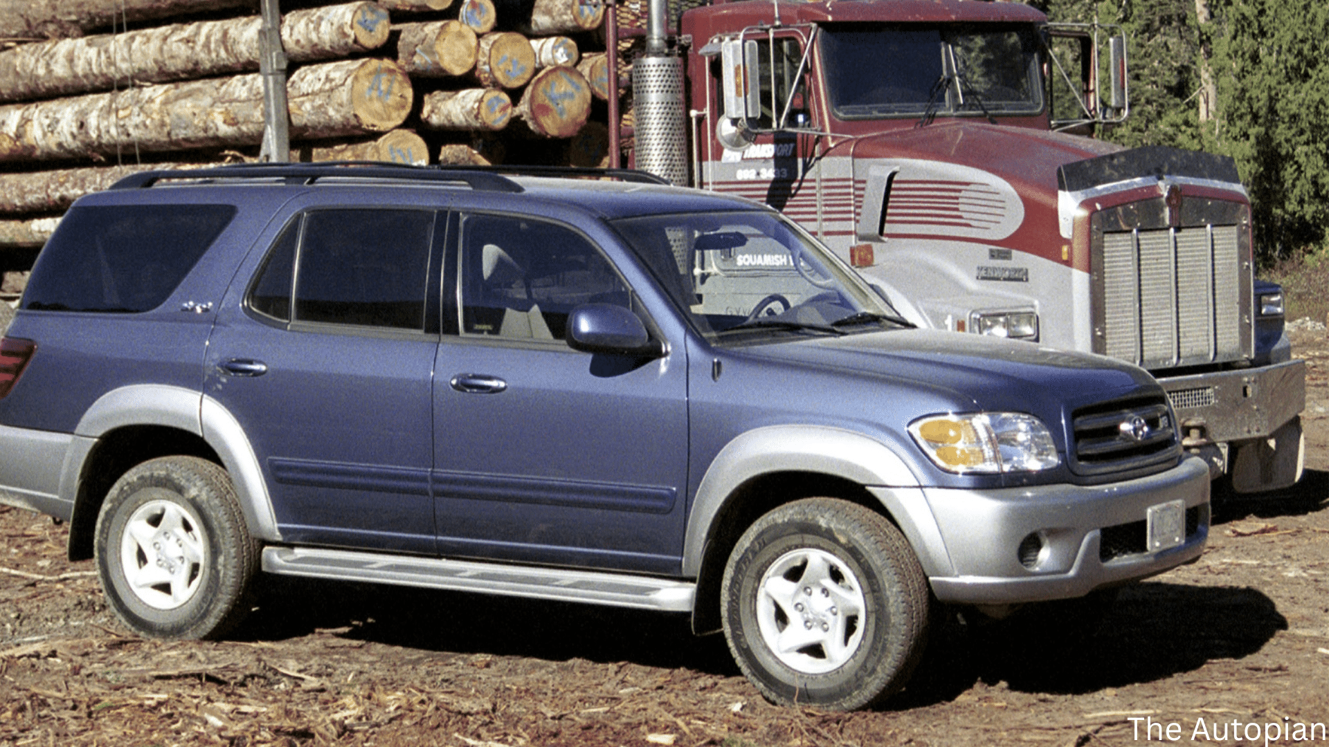 Blue Toyota SUV parked in a muddy logging yard beside a red semi truck loaded with large cut timber logs