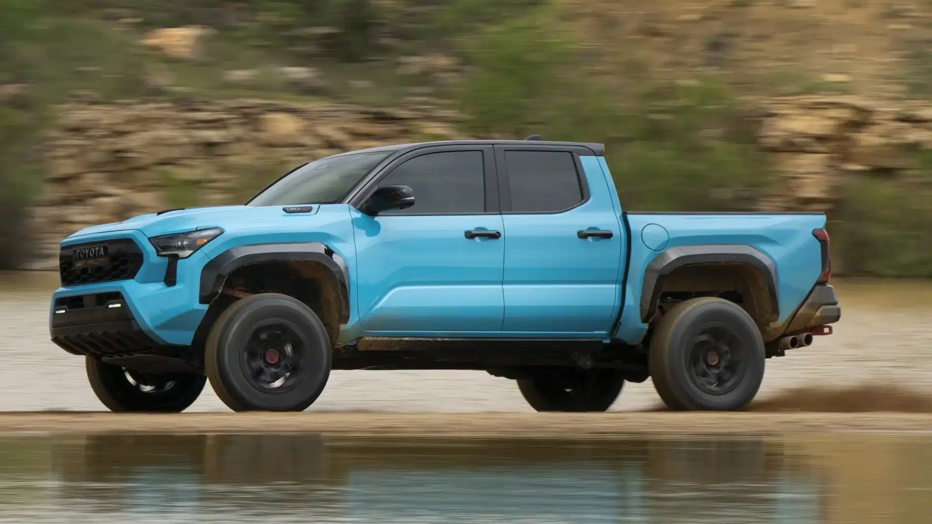 Blue Toyota Tacoma pickup driving beside water on rugged terrain during an off-road trail run in daylight