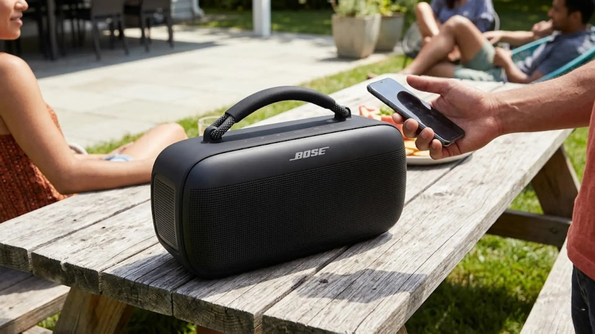 Bose portable speaker on outdoor table, person using phone, friends relaxing in backyard setting