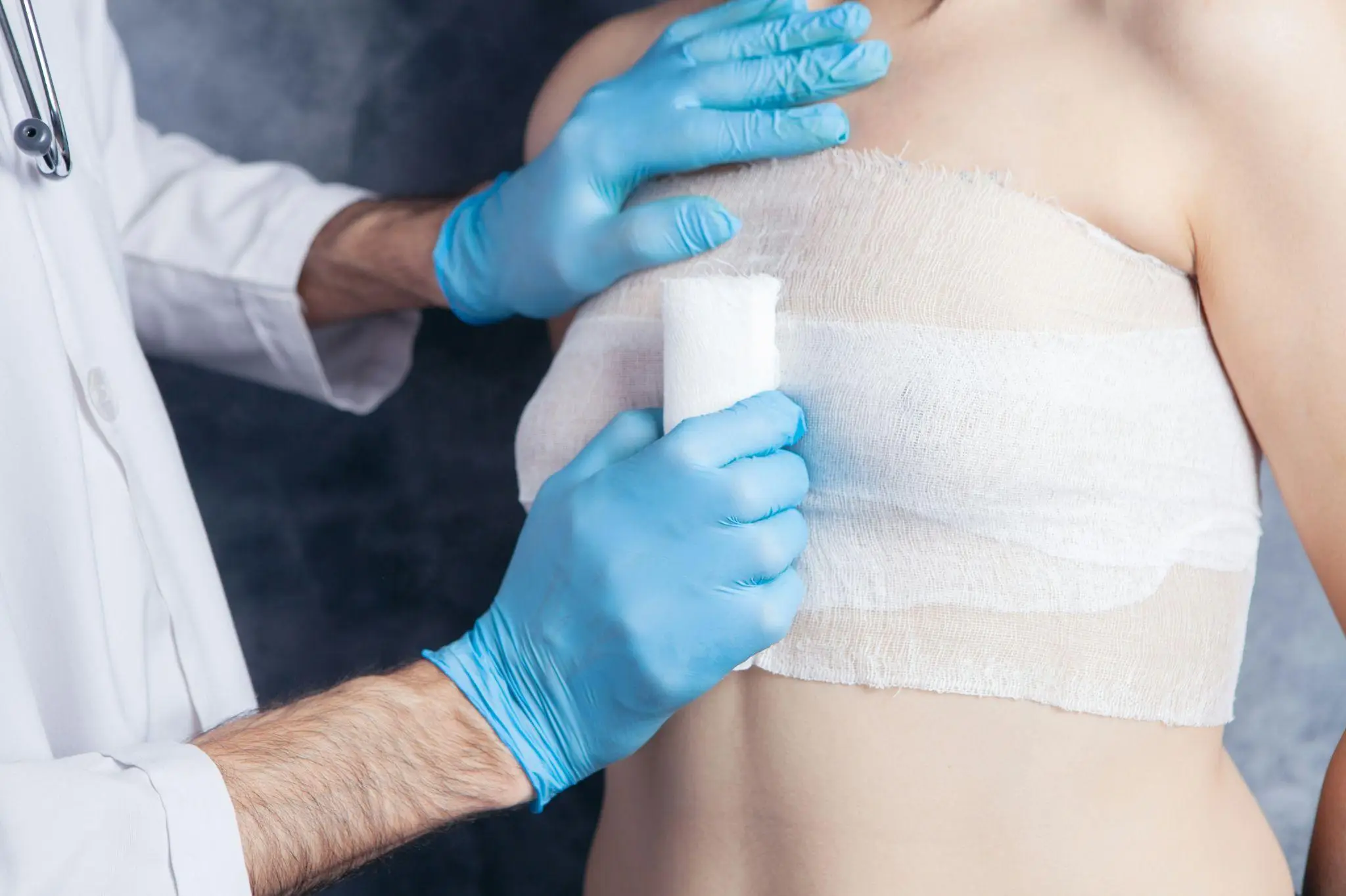 Medical professional applying bandage to patient’s chest wrapped in gauze, wearing blue gloves