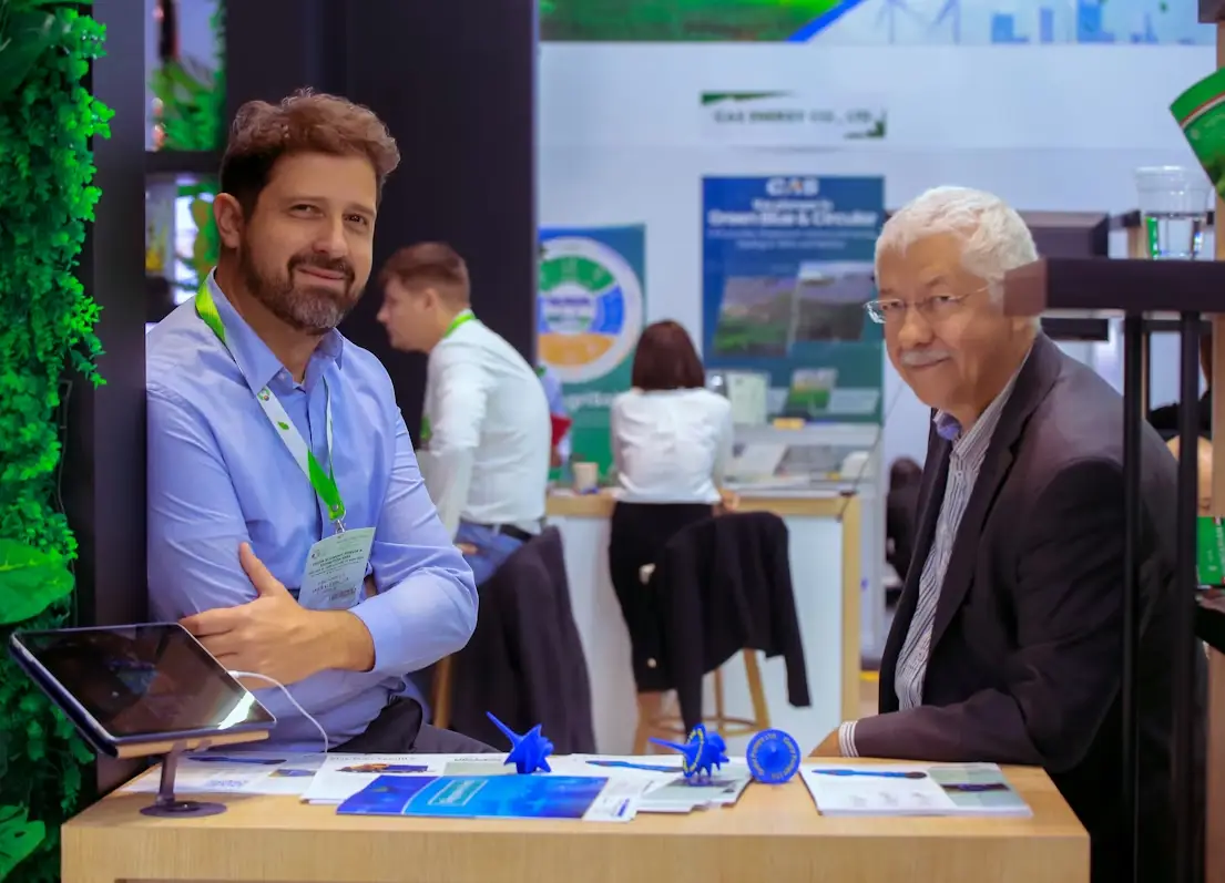 Two men sitting at a table with brochures and model turbines at a trade show