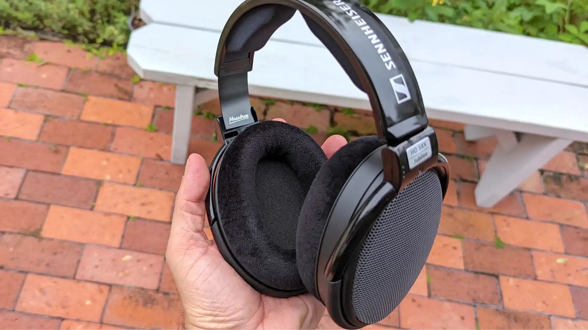 Hand holding black Sennheiser HD 58X Jubilee headphones outdoors near a white bench and brick patio.